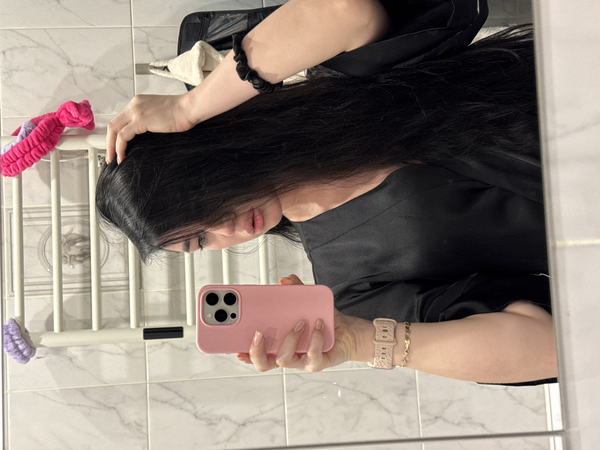 A young woman with long black hair is taking a mirror selfie in what appears to be a bathroom. She is wearing a black satin robe and a pink watch. In her right hand, she holds a pink smartphone, and her left hand is in her hair. A pink and purple scrunchie is visible hanging from a towel rack in the background. The bathroom walls are tiled with a marble pattern. The scene is set in Novosibirsk, Russia.