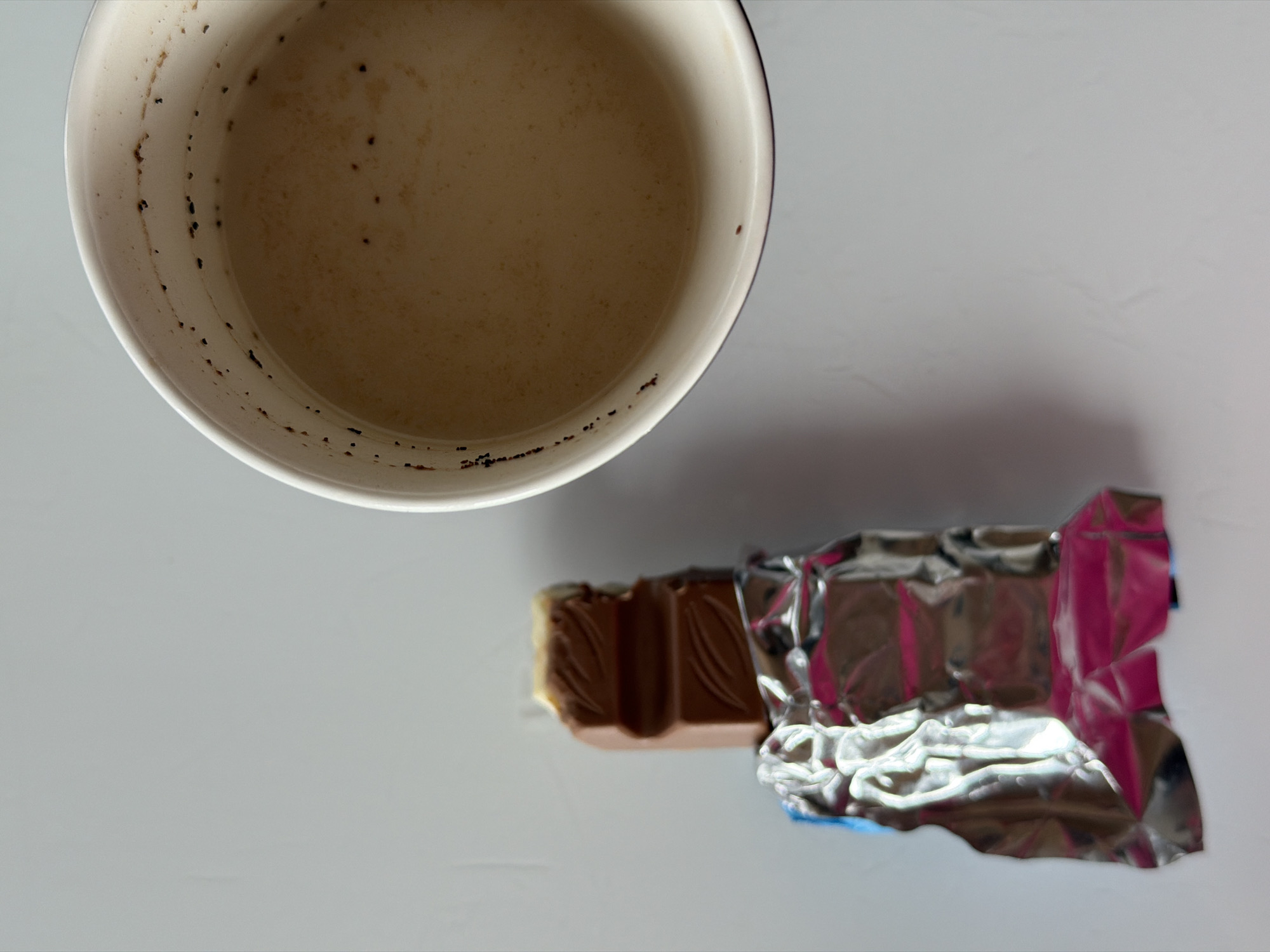 The image depicts a table with a partially consumed beverage in a white mug. The beverage appears light brown, possibly coffee with milk, with sediment visible along the inner rim of the mug. Next to the mug is a partially unwrapped chocolate bar. The chocolate bar is sectioned, with part of it exposed from its metallic wrapper. The table is a neutral color and the background is plain. The location of the image is unknown. The city and country specified are not visually discernible.