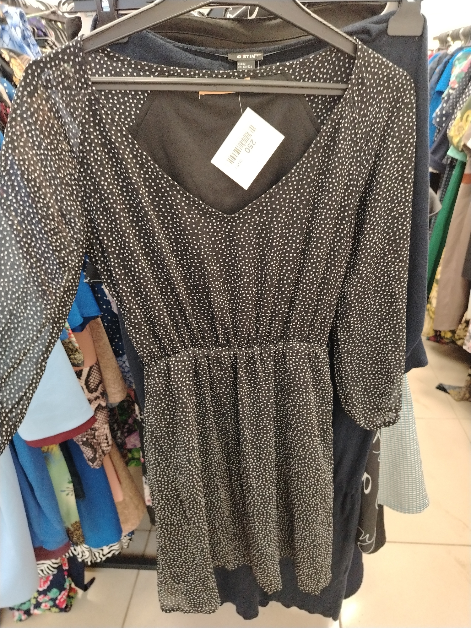 A black polka dot dress is displayed on a hanger in a clothing store. The dress has a V-neck and long sleeves. A price tag is attached to the dress. The background shows other clothing items and racks. The image was taken at O'STIN store in Yaroslavl, Russia.