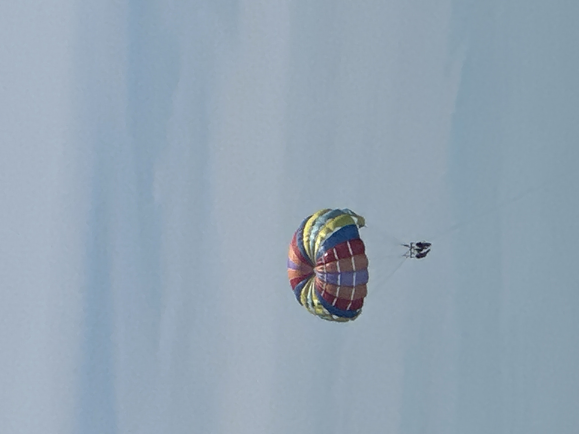 A colorful parasail, with panels of red, yellow, blue, and purple, is airborne against a pale blue, cloudy sky. Two figures are suspended beneath the parasail by harnesses and ropes, suggesting they are engaged in parasailing. The overall scene appears to be an outdoor recreational activity, likely taking place over water given the context of parasailing, though no water is visible in the frame. The location within the city of Boreti, Montenegro, is not specified by any visual cues in the image.
