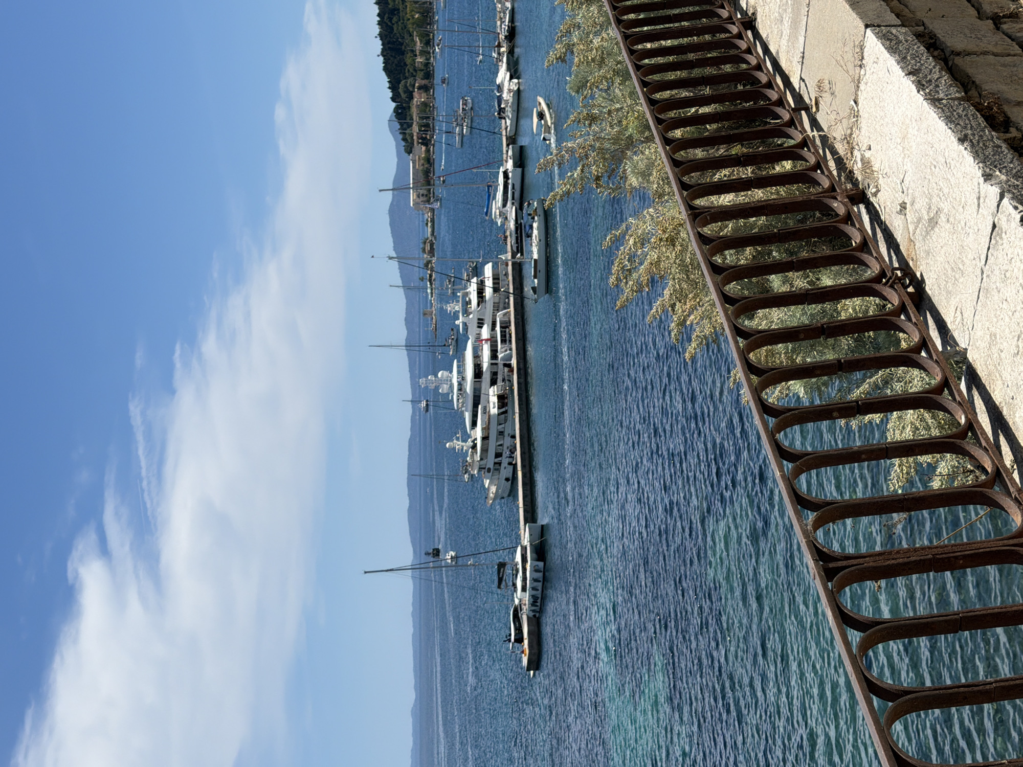 This image depicts a serene, sunlit marina scene in Corfu, Greece, under a clear blue sky with sparse white clouds. The vibrant blue-green waters of the Ionian Sea are calm and transparent, revealing the shallow seabed in places.
The scene is dominated by a collection of boats, ranging from large, gleaming white motor yachts and superyachts moored along a long pier, to smaller sailboats with their masts pointing skyward. Several boats are docked neatly, while others are visible further out in the bay, contributing to a picturesque port setting. In the background, distant hills and mountains, characteristic of the Greek landscape, rise gently, covered in sparse vegetation, adding depth to the horizon.
In the foreground on the right, a weathered and rusty metal railing with distinctive oval-shaped rungs angles downwards, bordering a robust, light-colored stone wall or embankment. Small patches of light green foliage grow around the wall and railing, suggesting natural overgrowth.
The image conveys a peaceful daytime atmosphere, indicative of pleasant weather suitable for maritime activities. The time of day is bright, with ample sunlight illuminating the entire scene. There are no people visibly present, allowing the natural and nautical elements to take center stage. No visible text is present in the image. The overall impression is that of a tranquil and beautiful Mediterranean coastal destination.