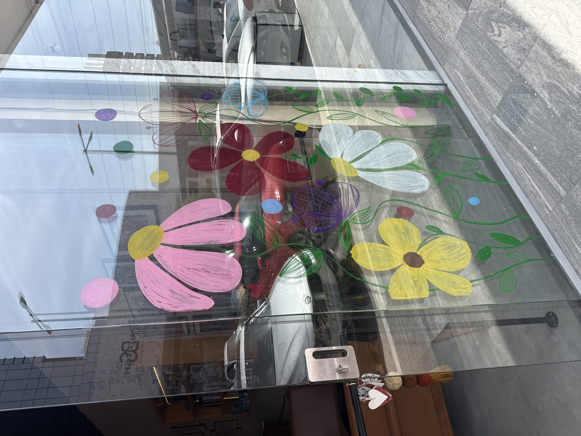 The image shows a window display in Puebla, Mexico, adorned with colorful floral paintings. The foreground reveals a glass pane decorated with a whimsical assortment of flowers in shades of pink, red, white, yellow, and purple. The blooms are accompanied by green vines and playful polka dots of various hues. The glass reflects the city scene outside, including buildings and a white car parked on the street. Through the window, a glimpse of the store's interior is visible, featuring shelves displaying various objects and a brown cabinet. A doorknob is seen at the bottom edge of the glass. The side of the building next to the storefront features a tiled facade.