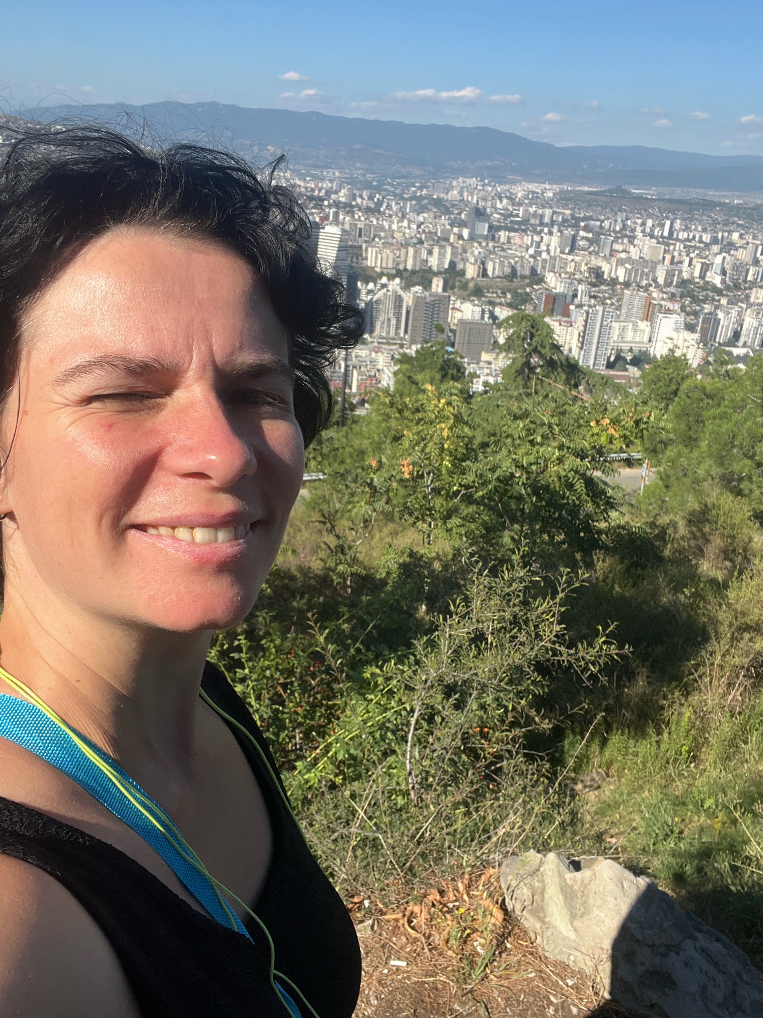 A woman with dark hair and fair skin takes a selfie outdoors. She is smiling and has one eye squinted. She is wearing a black tank top and a blue and yellow lanyard around her neck. Behind her, a sprawling cityscape with numerous buildings and mountains in the distance is visible. The scene appears to be a viewpoint overlooking Tbilisi, Georgia. The foreground includes green foliage and a large rock. The lighting suggests it is daytime, likely sunny.