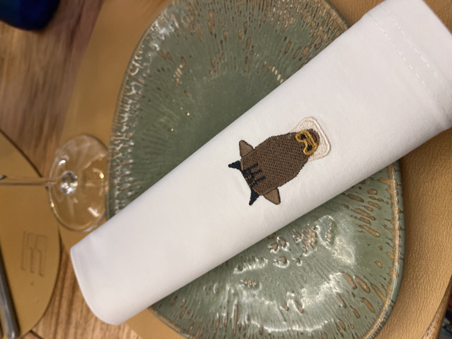 A white linen napkin with a brown bull embroidered on it is laying on a green ceramic plate, which is resting on a brown wooden table. There is a stemless wine glass, upside down, on the table to the left of the napkin. The coordinates 28.102, -15.414 correspond to the city of Las Palmas de Gran Canaria, Spain. The image is taken indoors, likely at a restaurant or café.