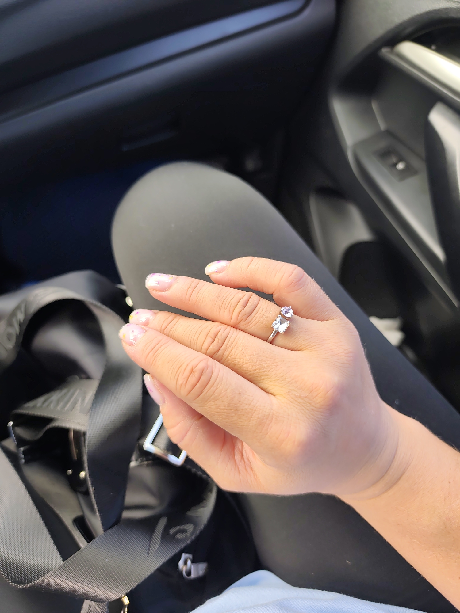 A person is seated in a vehicle, wearing black leggings. Their left hand is prominently displayed, adorned with a silver ring featuring two stones, one square and one round. The hand rests on a black bag with straps. The interior of the vehicle is visible, including the door panel and dashboard. The location, Chaikovsky, Russia, cannot be determined from the visual information provided.