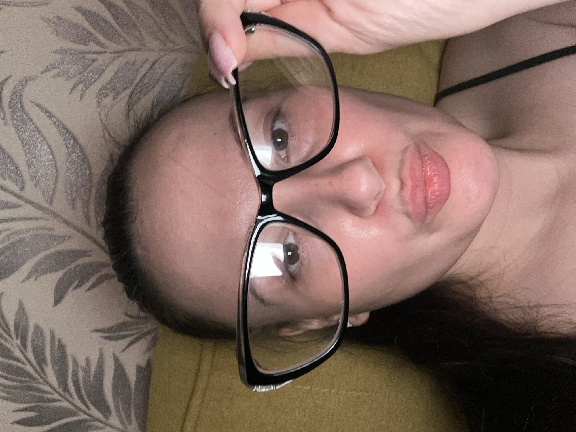 A woman with dark hair is lying on a light green and grey patterned surface, holding a pair of black framed glasses in front of her face. Her gaze is directed towards the viewer through the lenses. She has fair skin, a light pink lipstick, and partially visible black attire on her right shoulder. The background surface appears to be a cushion or upholstered piece of furniture. There is no information available to determine the location as Chaikovsky, Russia.