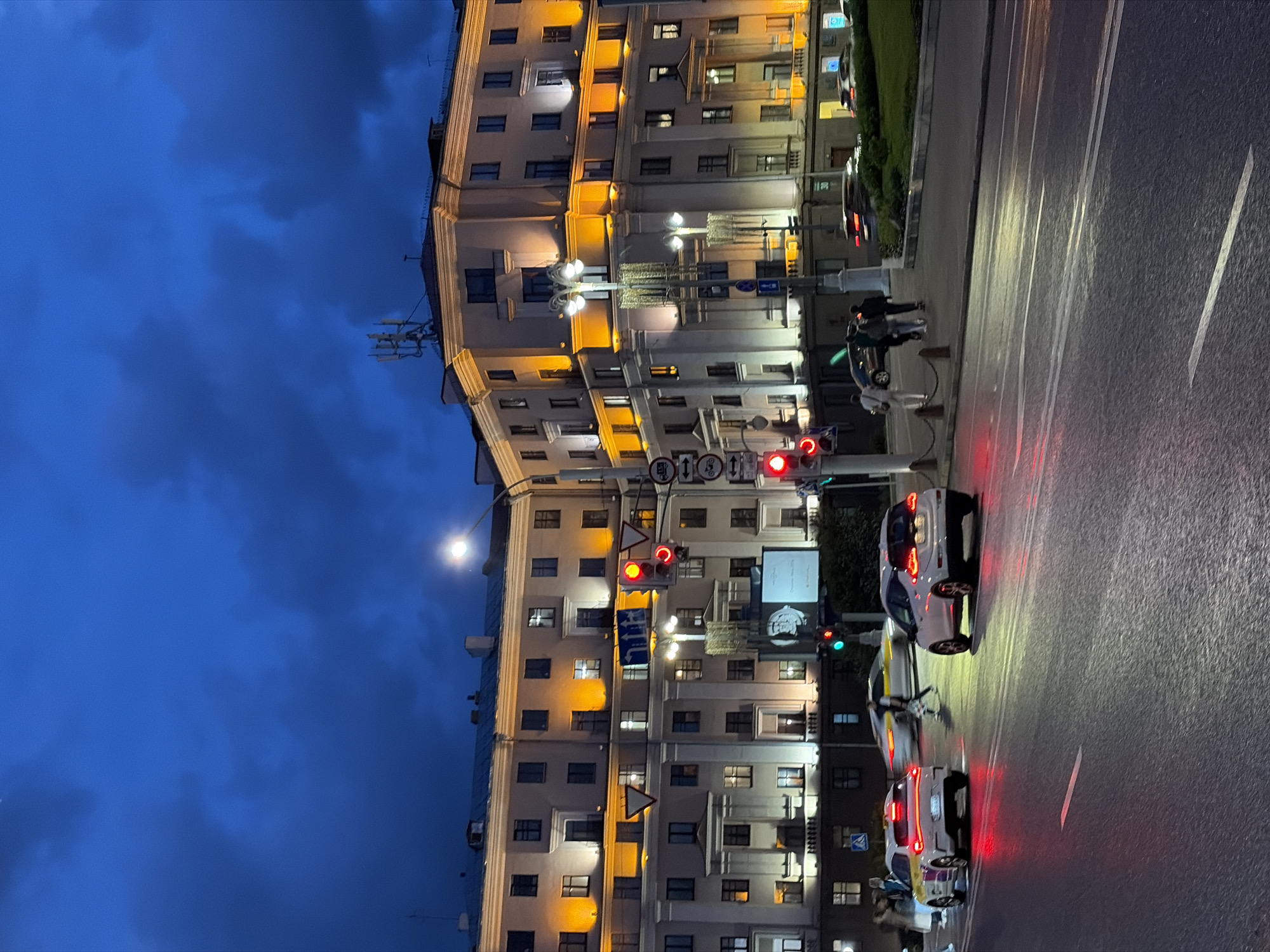 The image shows a street scene in Minsk, Belarus at dusk. A tall, light-colored building with multiple windows is illuminated with warm lights. A street runs along the right side of the building, with cars stopped at a traffic light. The traffic lights are red. Pedestrians are standing on the sidewalk near the street. The sky is a deep blue color with dark clouds. The street is wet, and the car headlights reflect off the pavement.