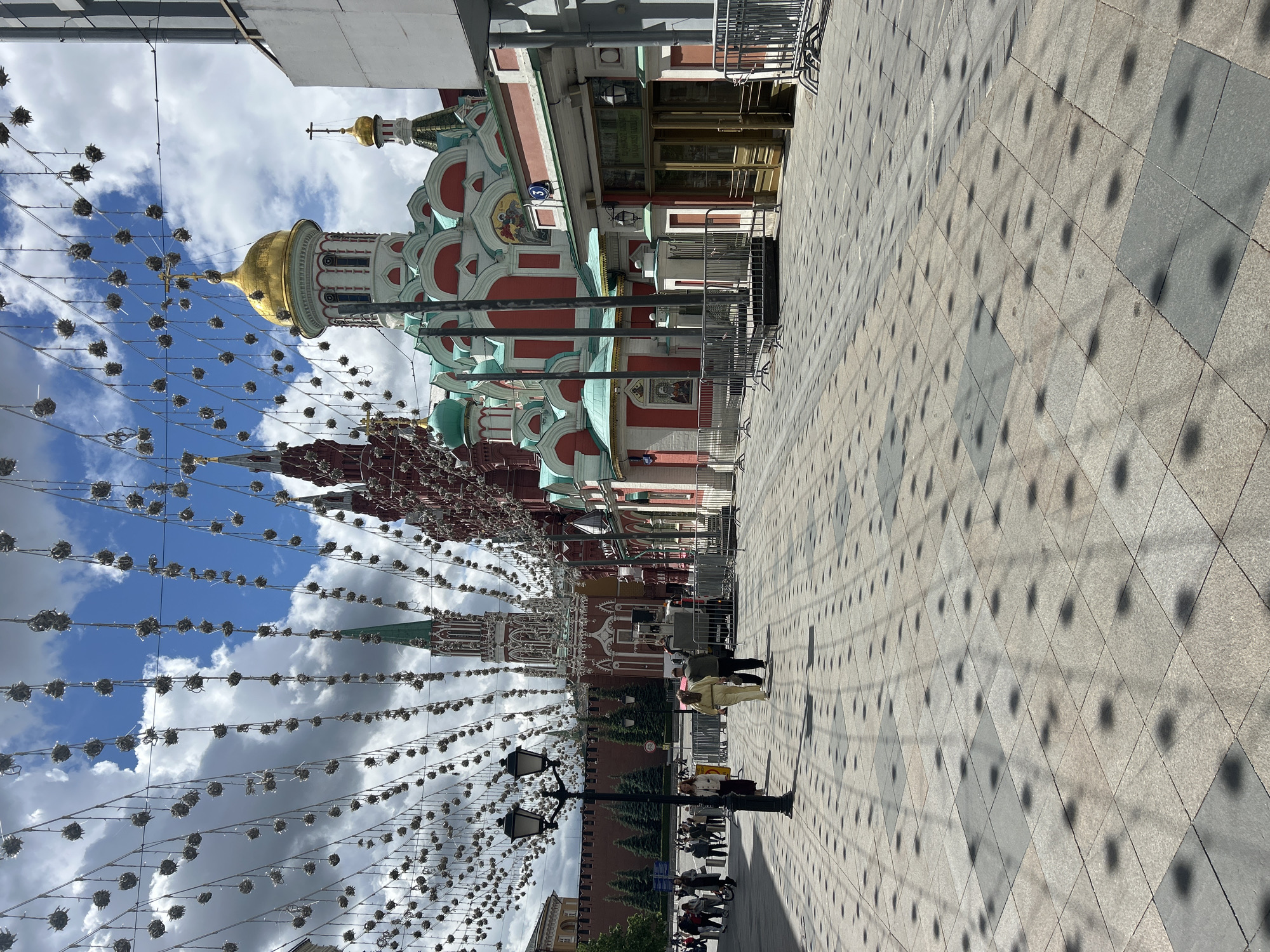 The image depicts a street scene in Moscow, Russia, showcasing architectural landmarks. The foreground features a paved pedestrian area with visible shadows from overhead decorations. A colorful building with red and white facades and golden domes is prominently displayed, which is recognizable as a church. Beyond the church, part of the red walls and towers of the Kremlin are visible. Decorative strings of lights and ornaments are strung across the street. There are people visible, mostly in the middle distance. The sky above is partly cloudy. A streetlamp is also apparent in the shot.