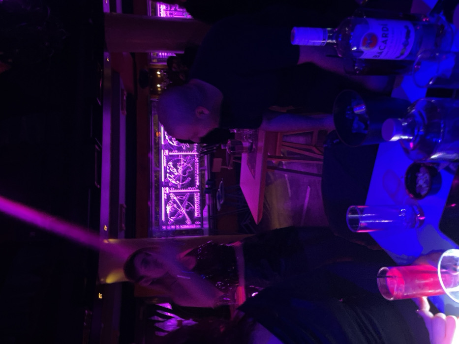 This media file captures an indoor scene at a nightlife venue, likely a bar or club, characterized by atmospheric low light and vibrant purple and blue illumination. The image, originally rotated, shows a dynamic setting during the nighttime.
In the foreground and midground, a table is visible, adorned with several glasses and bottles. Prominently displayed is a bottle of "BACARDI" rum. One glass on the table appears to hold a red beverage.
Two individuals are partially visible. On the left side of the frame, a person with a bald or closely shaven head, wearing a dark top, leans over the table. On the right, another person with dark hair is seen, partially blurred, looking towards the right side of the frame. Their expressions are indistinct due to the dim lighting.
The background features decorative panels brightly lit with purple, showcasing intricate, stylized designs or figures, enhancing the venue's artistic ambiance. A distinct beam of purple light cuts across the upper left portion of the image, contributing to the club-like atmosphere. The overall scene suggests a social gathering where people are enjoying drinks and the vibrant environment of a bar in Drapetsona, Greece.