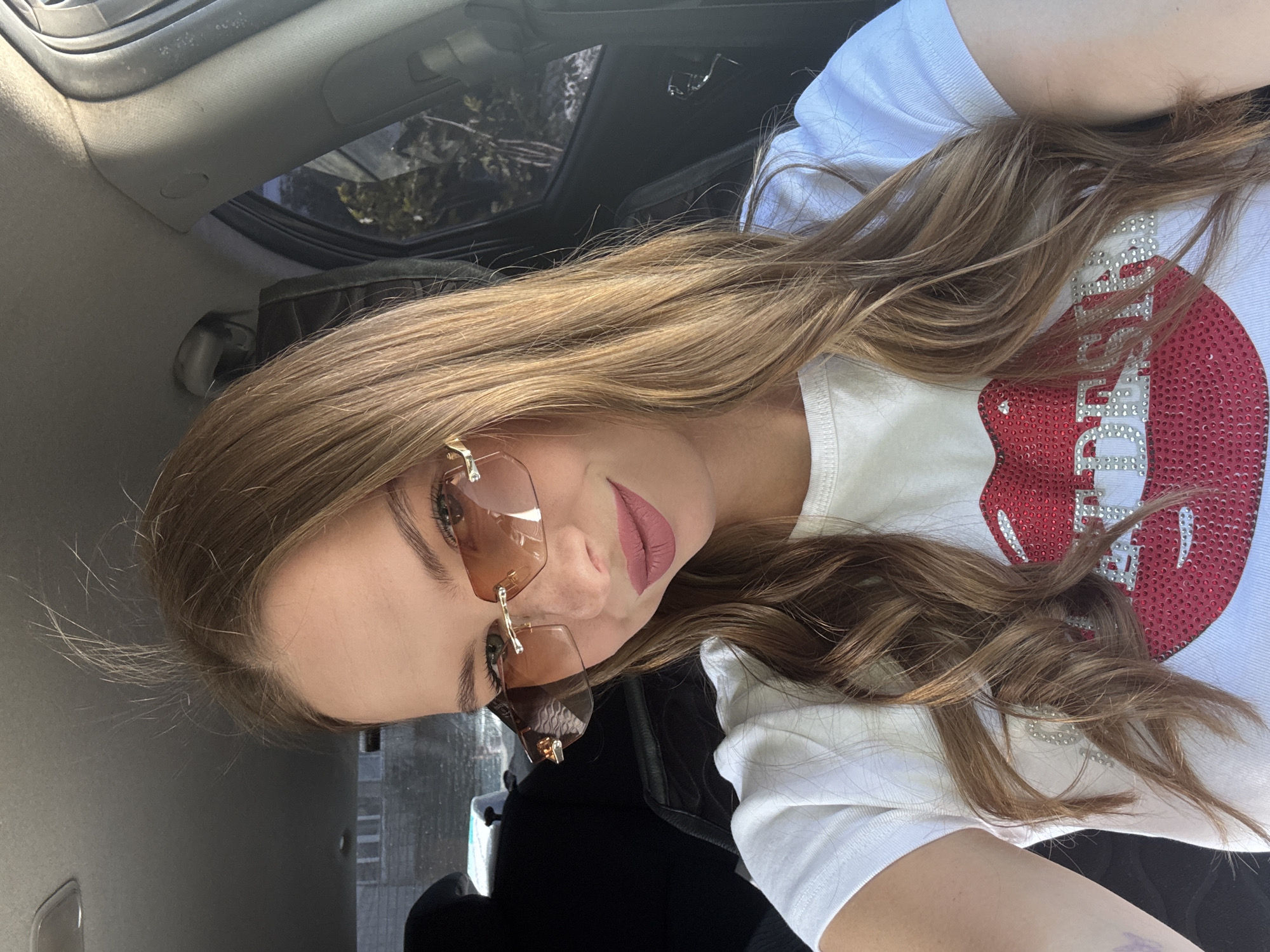 A young woman with long, blonde-brown hair is sitting in the driver's seat of a car, taking a selfie. She is wearing oversized, rimless sunglasses with a gradient brown lens and a white t-shirt adorned with a large, sequined red lip design and silver lettering. Her lips are painted a matte pink color. The interior of the car is visible, including the dashboard, a black car seat, and the car's headliner. Through the car window, a building with visible windows and a cloudy sky can be seen. The overall lighting suggests it is daytime.