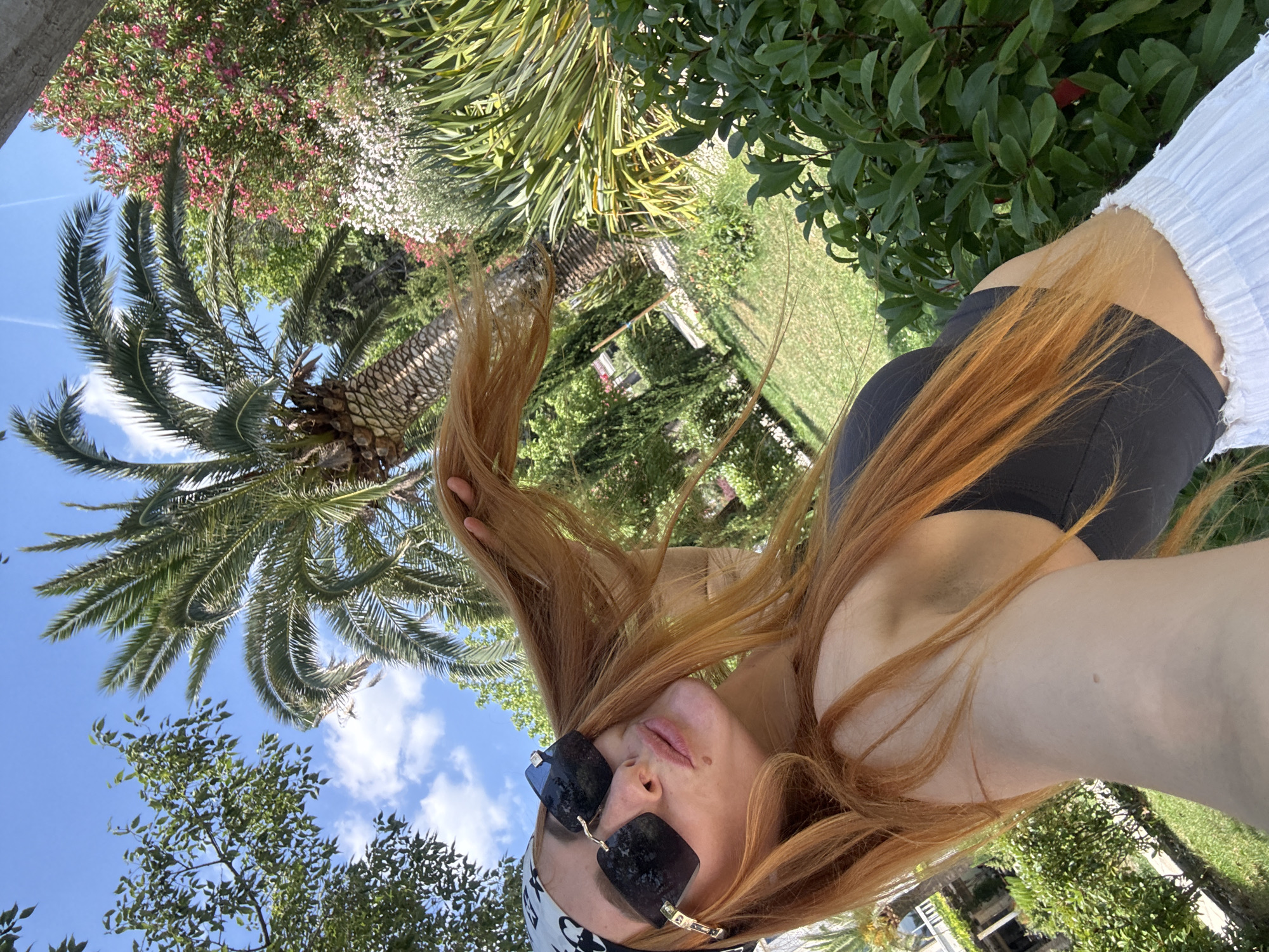 The image is a low-angle, vertical selfie of a young woman with long, reddish-blonde hair. She is wearing large, square black sunglasses with gold accents, and a white headband with a black pattern. Her hair is flowing around her face and shoulders. She is wearing a black crop top and a white ruffled skirt or cover-up. She appears to be outdoors in a sunny, tropical or Mediterranean-like setting. Lush green foliage, including palm trees and flowering bushes with pink and white blossoms, dominates the background. A bright blue sky with scattered white clouds is visible above. The overall impression is one of a relaxed, summery day. The lighting suggests it is daytime, likely late morning or early afternoon. The scene appears to be a garden or a resort area, with hints of lawn furniture in the background. The image evokes feelings of warmth and leisure. No visible text is present.
