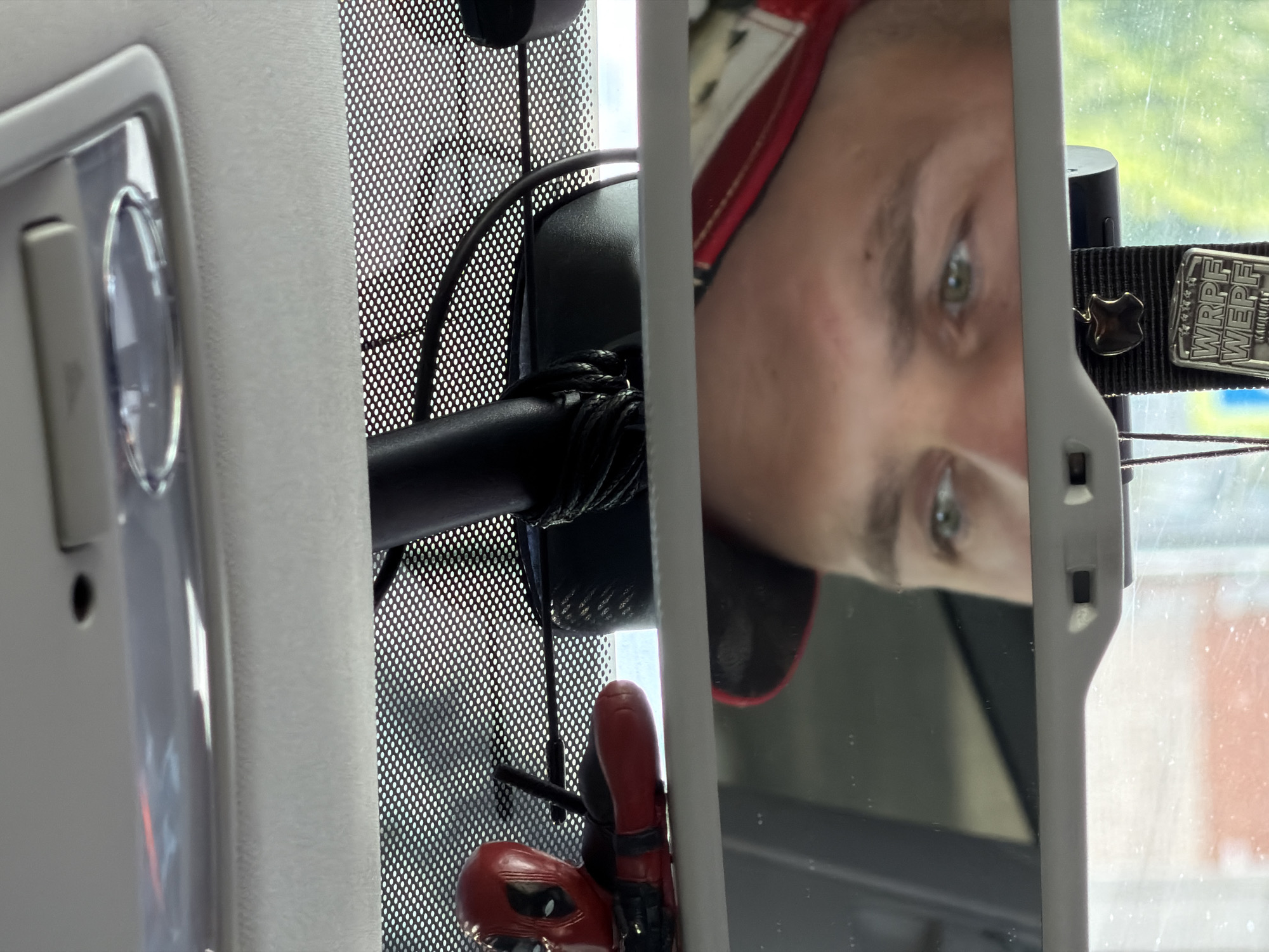 The image appears to be a reflection in a car's rearview mirror. The reflection shows a person's face with light-colored eyes. Attached to the rearview mirror is a black strap with a metal pin and an enameled emblem labeled "WRPF WEPT." Hanging from the mirror support is a small Deadpool figurine. The background suggests a car interior with a mesh sunshade on the windshield. Given the prompt, the implied location is Chaikovsky, Russia, but this cannot be confirmed from the image itself.