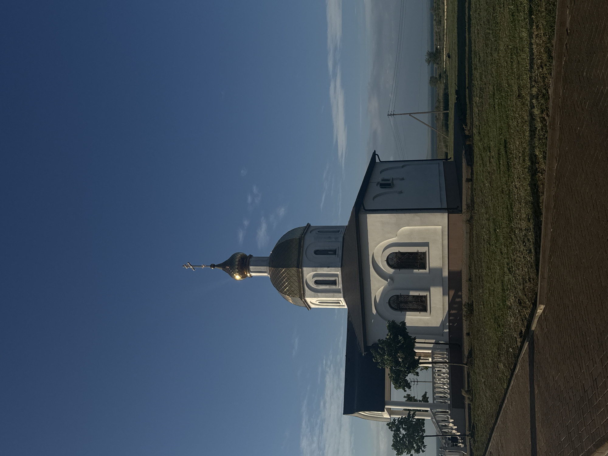 The image shows the Church of the Intercession of the Holy Virgin in Taman', Russia. The church has a golden dome and a cross on top. The building is white with arched windows. The image is taken from a low angle, looking up at the church against a clear blue sky. A patch of green grass and part of a brick sidewalk can be seen on the right. There are trees in the front of the church. Powerlines extend from the background.