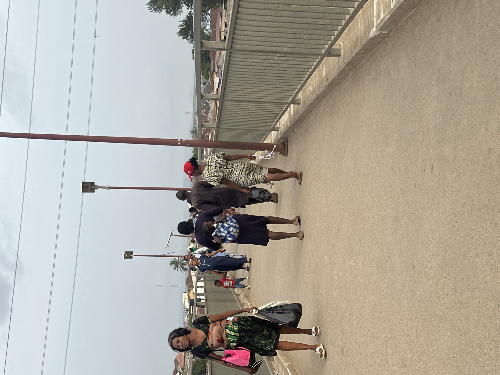 The image shows a group of people walking on a pedestrian bridge in Kuchingoro, Nigeria. The bridge has a concrete surface and metal railings. Utility poles are visible along the side of the bridge. The people include women, men, and children. Some individuals are carrying bags or objects. The background shows buildings and vegetation, typical of an urban environment. The sky is overcast.