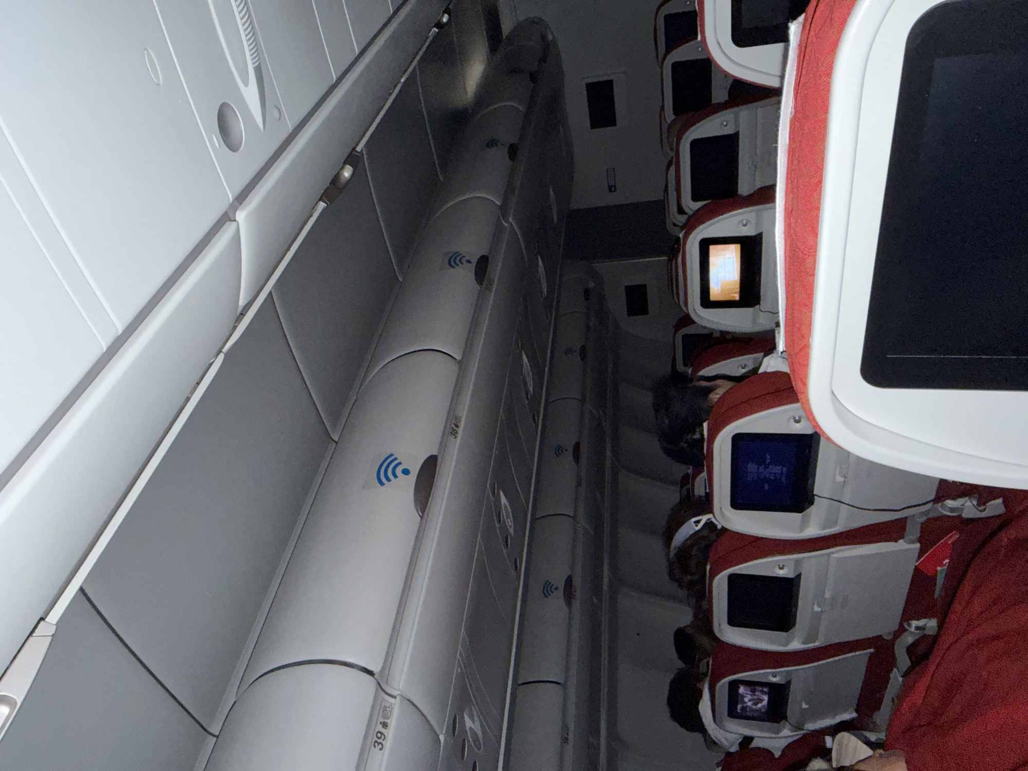 An interior view of an airplane cabin, likely an economy class section, is captured with a dim, ambient lighting, suggesting a night flight or relaxed cabin atmosphere. The image is oriented vertically, showing the length of the cabin from a perspective near the window or aisle, looking towards the opposite side.
On the right side of the frame, several rows of aircraft seats are visible. The seats feature red fabric upholstery and white or light-colored frames, each equipped with a personal entertainment screen on the seatback. Some screens display content, including one with illuminated text or an image, while others are dark or off. Passengers are seated, with their heads and shoulders partially visible, appearing to be resting or engaged with the in-flight entertainment.
The left side of the frame showcases the overhead bins and ceiling panels of the aircraft. These are predominantly white or light grey. Multiple blue Wi-Fi symbols, depicting three curved lines radiating upwards, are affixed to the underside of the overhead bins, indicating the availability of in-flight internet connectivity. One overhead bin clearly shows the seat number "39 A". The overall impression is a typical commercial flight cabin during a period of low activity, with passengers settled in their seats. The provided location context, Mysy, Russia, is not directly discernible from the interior of the aircraft.