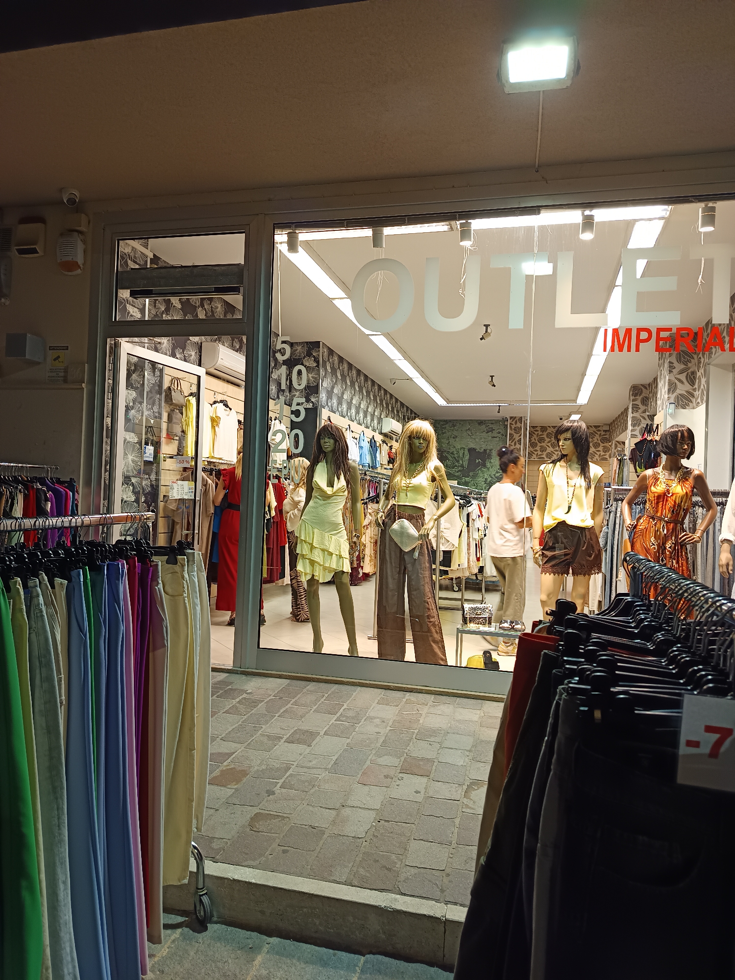 This image depicts the storefront of "OUTLET IMPERIAL" located on the Corso Primo Maggio in Roseto degli Abruzzi, Italy. The shop features a large display window showcasing mannequins dressed in various outfits, illuminated by interior lighting. Outside the window, clothing racks with an array of colorful garments are visible. The street in front of the store is paved with cobblestones. A security camera is mounted on the exterior wall of the building.