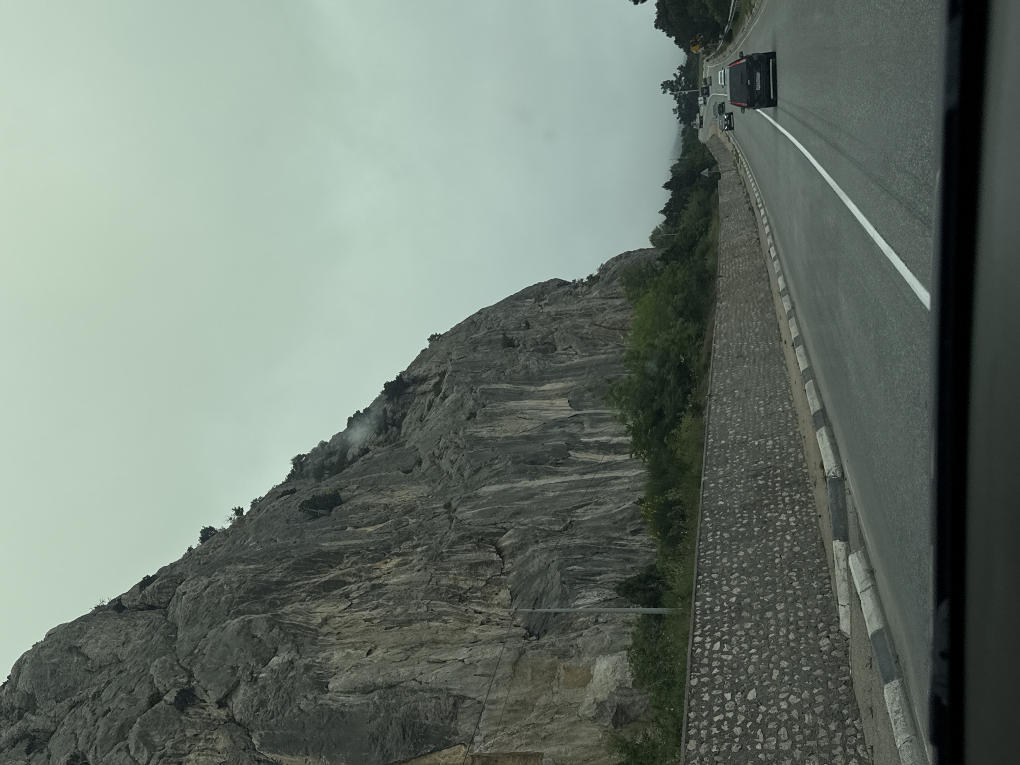 The image shows a road running alongside a rocky mountain. A line of vehicles is traveling on the road. The sky is overcast. The location appears to be a mountainous area. Details of a city are indiscernible.