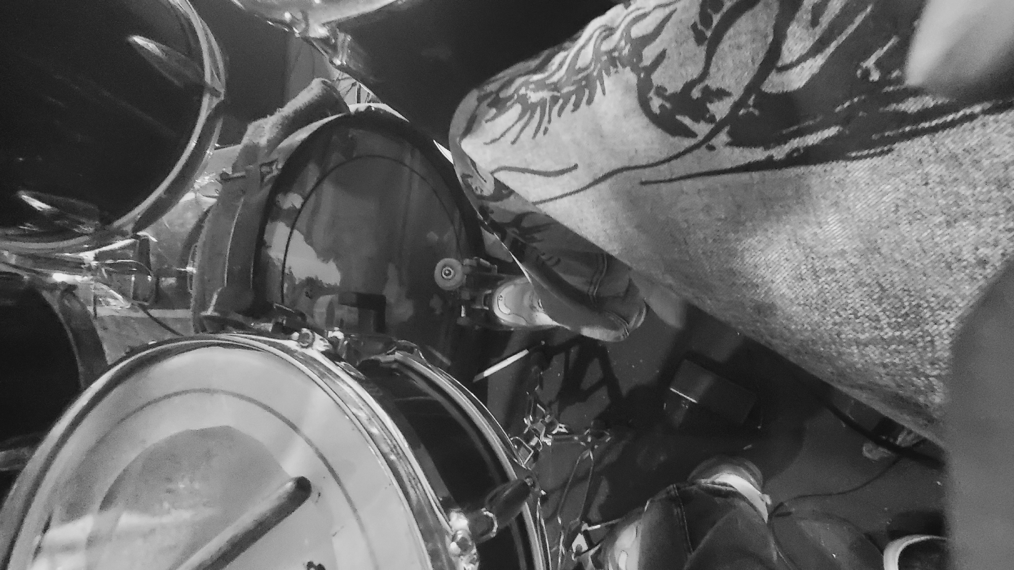 This is a black and white, high-angle, close-up shot of a person playing a drum kit. The perspective is from the drummer's point of view, looking down at their legs and the drums. The focus is on the drummer's left leg, clad in patterned jeans, and their foot on the bass drum pedal. A drumstick is visible resting on the snare drum. The scene is indoors, as suggested by the presence of musical equipment. There is no visual information to determine the specific location within Jalingo, Nigeria.