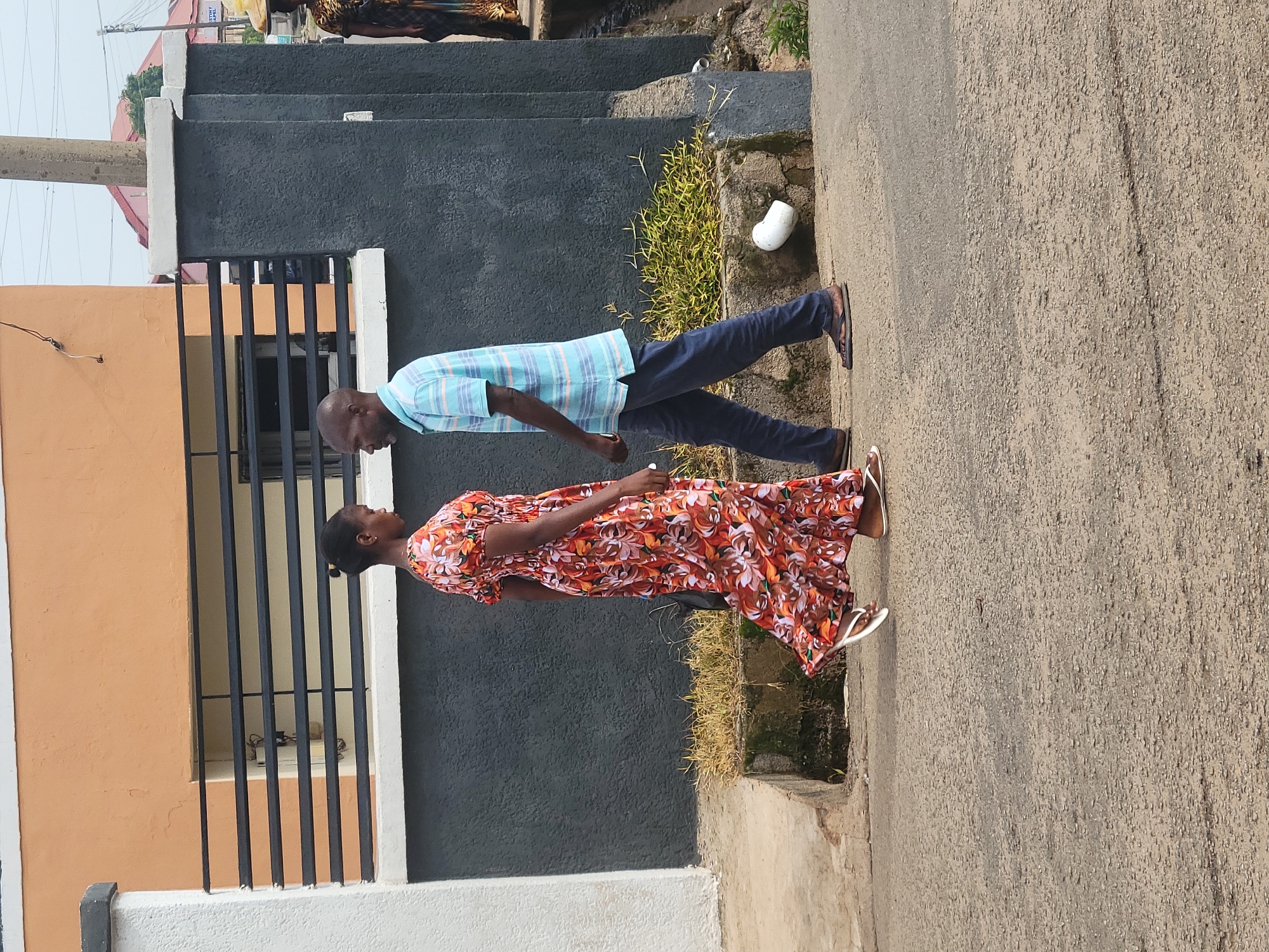A man and a woman are walking on a paved street in Jos, Nigeria, facing each other. The woman, on the left, is wearing a long, orange, floral-patterned dress and white flip-flops. She has her hair pulled back in a bun. The man, on the right, is wearing a light blue and white striped short-sleeved shirt, dark blue trousers, and brown sandals. They are positioned in the middle ground of the image, with a building behind them and the street in the foreground. The building has an orange upper section with black horizontal bars across windows, and a dark gray lower section. A concrete fence with white trim separates the building from the street. To the far right, the edge of another building and a utility pole are visible. The ground appears to be a street or pathway, with some overgrown vegetation and a white pipe fragment visible near the base of the fence. The sky is overcast.
