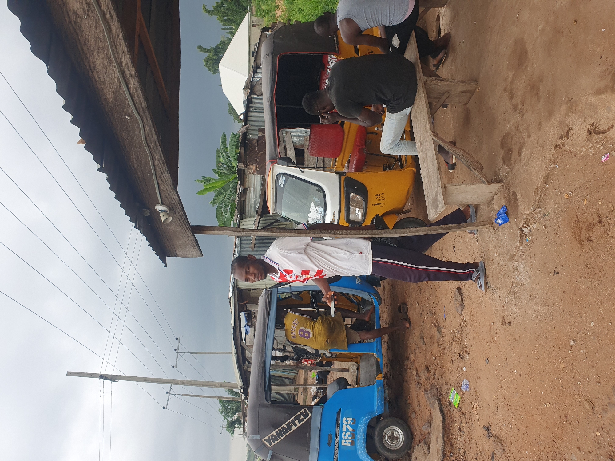 The image captures a scene in Lokoja, Nigeria, where three auto-rickshaws, also known as "keke napep" or "marwa", are parked. Two of the vehicles are blue and yellow, while a third is partially visible. A wooden bench sits in the foreground, occupied by two men. One man, wearing a black t-shirt and light-colored jeans, is looking down, possibly at his phone. Beside him, another man in a grey tank top and dark shorts is also seated, his back to the camera.
Standing near the blue auto-rickshaw is a man in a white and red striped shirt and dark pants, holding a phone in his hand and looking towards the camera. Another man, wearing a yellow jersey with the number "8" and the name "Bryant" on it, is partially visible inside the blue auto-rickshaw, his legs bent as he sits.
The environment appears to be a dusty, open area with some makeshift structures and corrugated metal roofing visible in the background. Utility poles and power lines are prominent in the sky, suggesting an urban or semi-urban setting. The ground is covered in red-brown soil and scattered debris. The overall impression is a typical roadside scene in a Nigerian city.