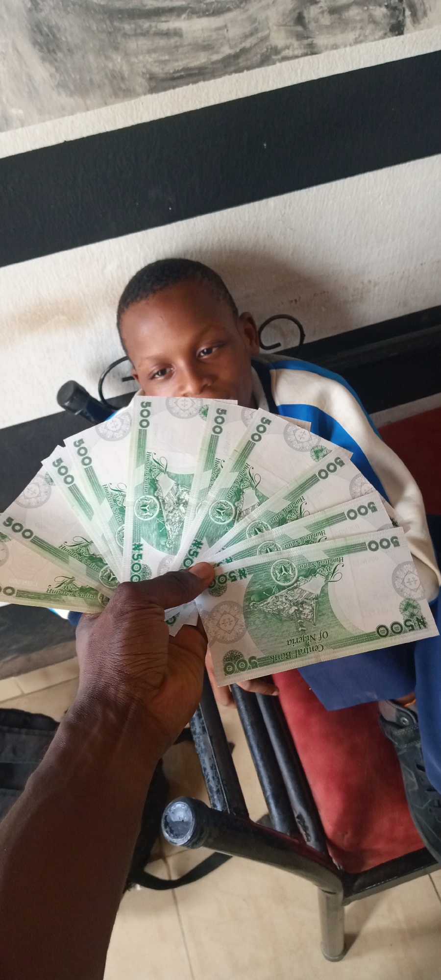 A medium-angle shot depicts a young boy seated, partially obscured by a fan of Nigerian Naira 500 notes held in the foreground by a darker-skinned hand. The boy has short, dark hair and is wearing a jacket with blue and white stripes. He is sitting in what appears to be a chair with a red cushion and black metal frame.
The hand holding the money is positioned centrally in the lower half of the image. The fan of banknotes prominently features the "500" denomination and the words "Central Bank of Nigeria."
The background consists of a wall painted in alternating bands of white and black. The location is identified only as "Unknown, Unknown."