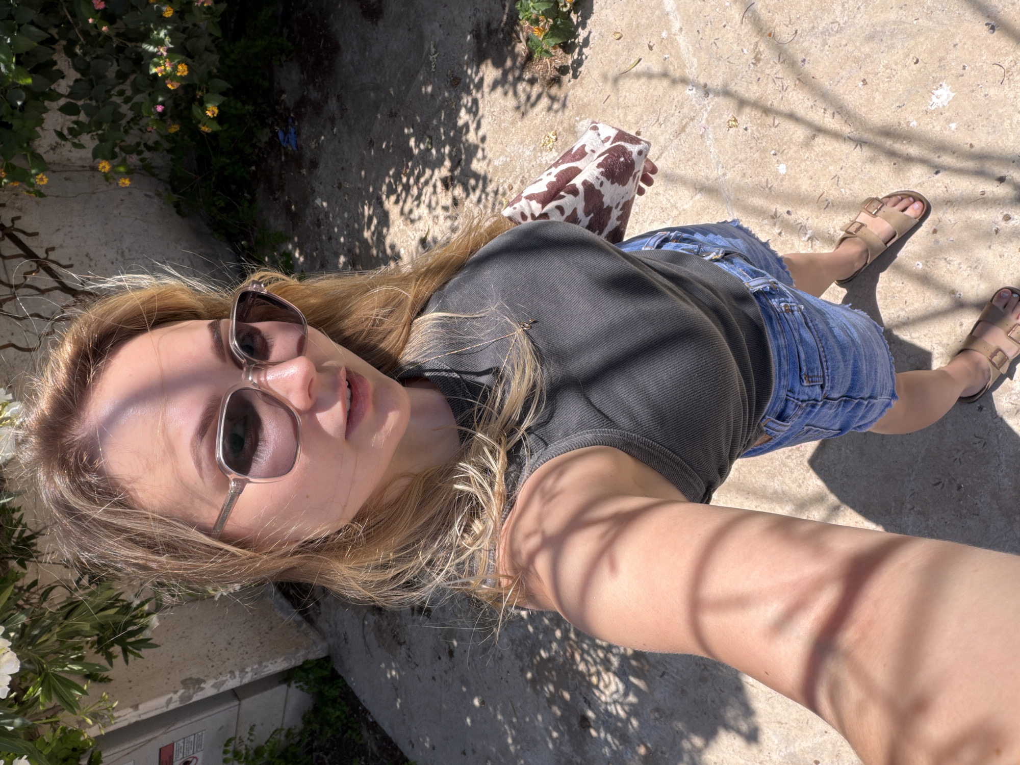 The image is a bright, sunny outdoor selfie featuring a young woman. The composition is rotated 90 degrees clockwise, showing the woman positioned diagonally, looking upwards towards the camera.
**Subjects**: The main subject is a young woman with long, blonde to light brown hair. She is wearing stylish sunglasses that reflect the bright sunlight. Her expression is relaxed, with a slight smile. She is dressed in a dark gray or black ribbed tank top, light blue denim cutoff shorts, and light brown, two-strap sandals. A thin necklace is faintly visible on her chest. In her right hand, near her waist, she holds a distinctive accessory or small bag with a brown and white cow-print pattern.
**Scene and Setting**: The scene is outdoors on a clear, sunny day, likely in Tel-Aviv, Israel, as per the context. The ground beneath her is concrete, covered in strong, dappled shadows cast by overhead foliage, suggesting trees or bushes. In the background and edges of the frame, green plants with small yellow and pink flowers are visible, indicating a garden or a landscaped outdoor area. A concrete wall or planter box is also visible.
**Activity**: The woman is actively taking a selfie, holding the camera at a low angle and looking up towards it.
**Notable details**: The prominent shadows and bright highlights on her face and clothing confirm a sunny day, likely during mid-day or afternoon. The overall mood is casual and summery. There is no discernible text visible in the image.