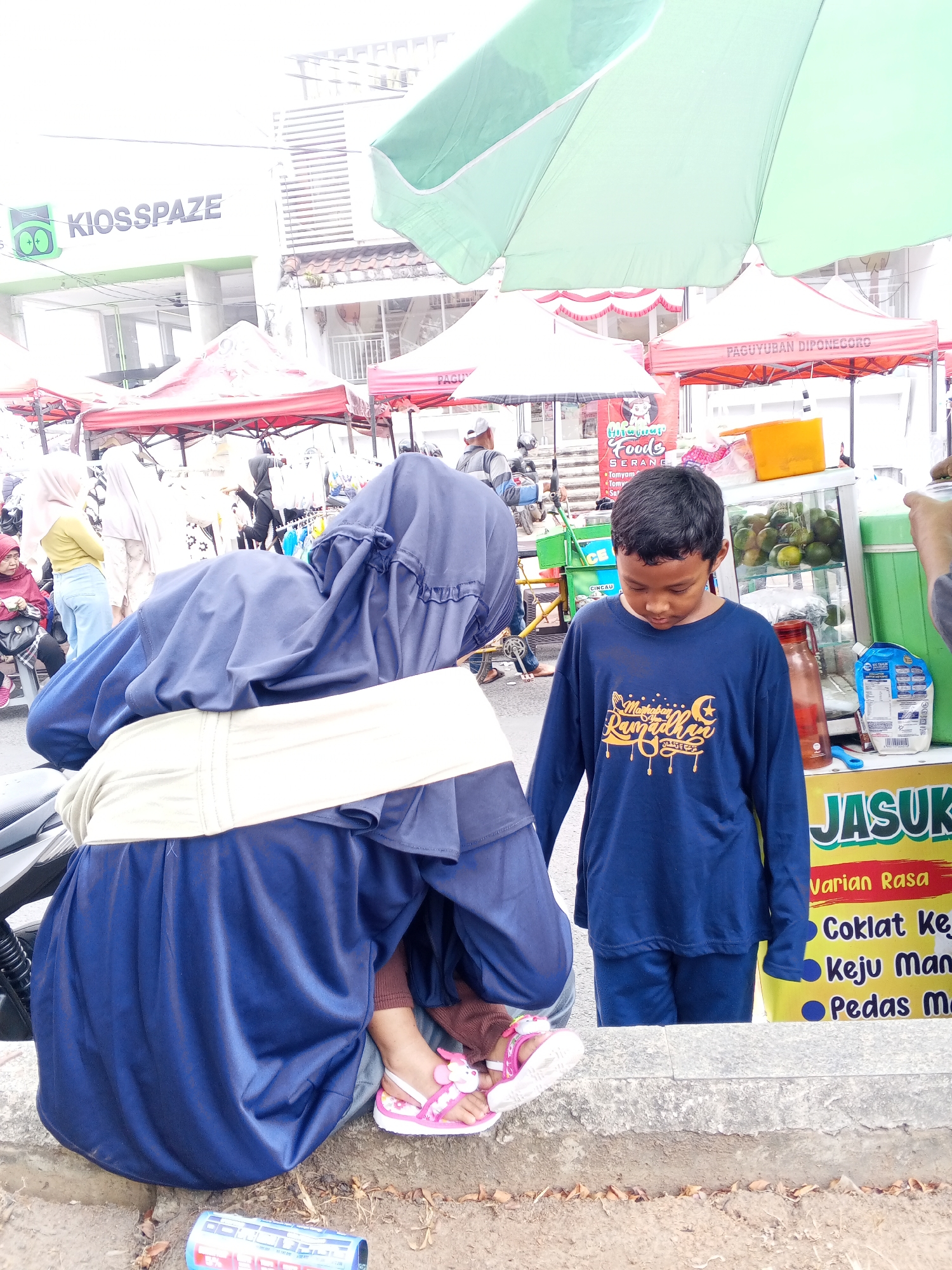 The image depicts a street scene in Serang, Indonesia, likely a market or roadside stall area. In the foreground, a person wearing a dark blue hijab and clothing is crouched down, with their back to the camera. They are wearing pink sandals with a cartoon character on them. Next to them stands a young boy in a dark blue long-sleeved shirt with yellow writing that appears to say "Marhaban Ramadhan". Behind them, a food stall is visible with a yellow sign advertising "JASUK". The sign lists "Varian Rasa" (Flavor Variants) including "Coklat Keju" (Chocolate Cheese), "Keju Manis" (Sweet Cheese), and "Pedas Manis" (Sweet Spicy). The stall also displays what appear to be oranges or limes in a glass case and various drinks. In the background, there are other market stalls with red and white awnings, as well as a building with the sign "KIOS SPAZE". The overall environment suggests a lively outdoor commercial area.