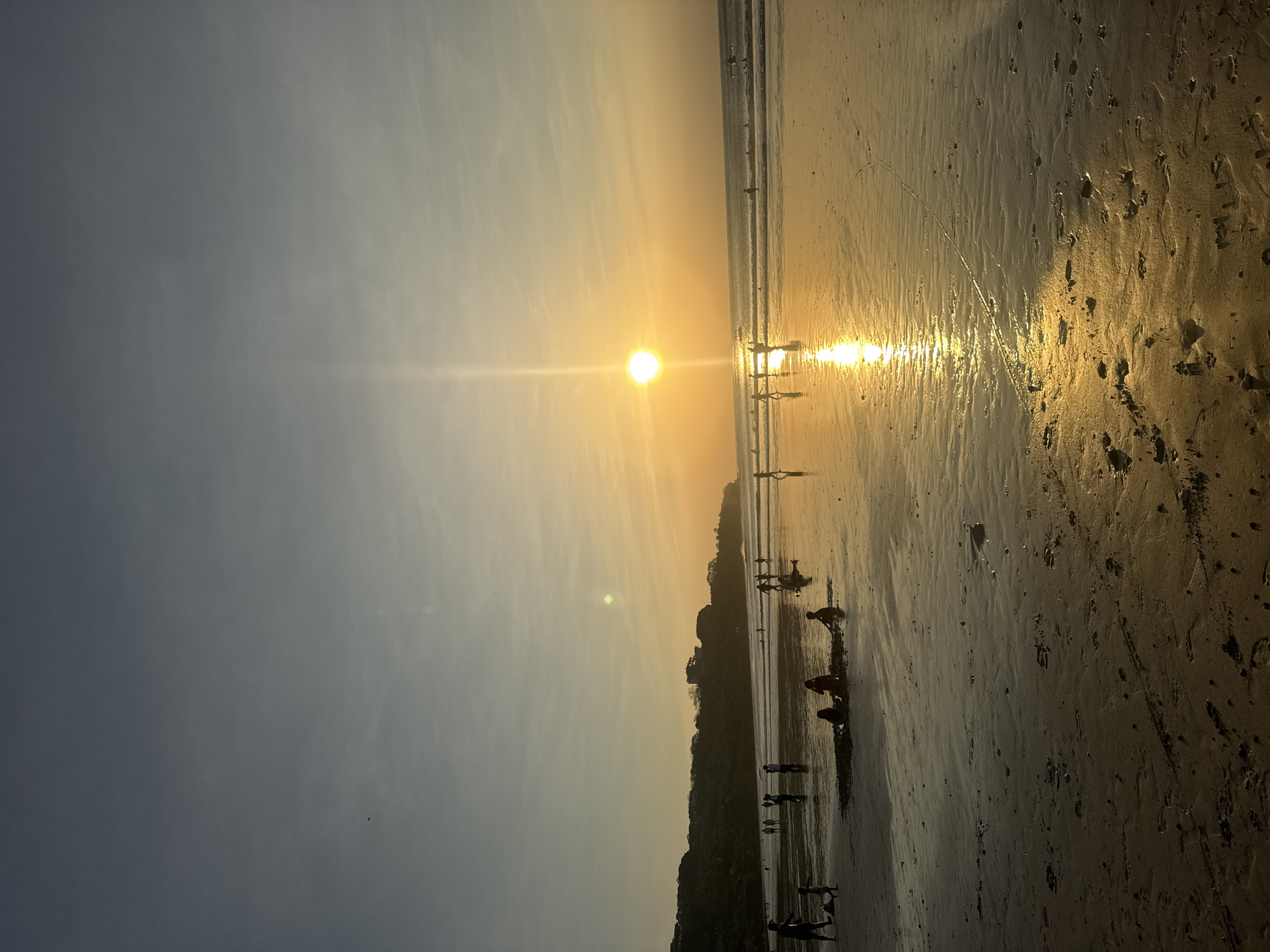 At sunset on Kuta Selatan beach, a body of water reflects the golden hues of the sky, casting a shimmering light across the wet sand. Silhouettes of people are visible across the expanse of the beach, some wading in the shallow water, others walking along the shore, and a few gathered in small groups on the sand. The sun, a bright orb of light, is low on the horizon, its rays extending upwards and illuminating the scene with a warm glow. The background shows a lush, tree-covered hillside with buildings dotting its slopes. The overall atmosphere is one of peaceful evening activity.