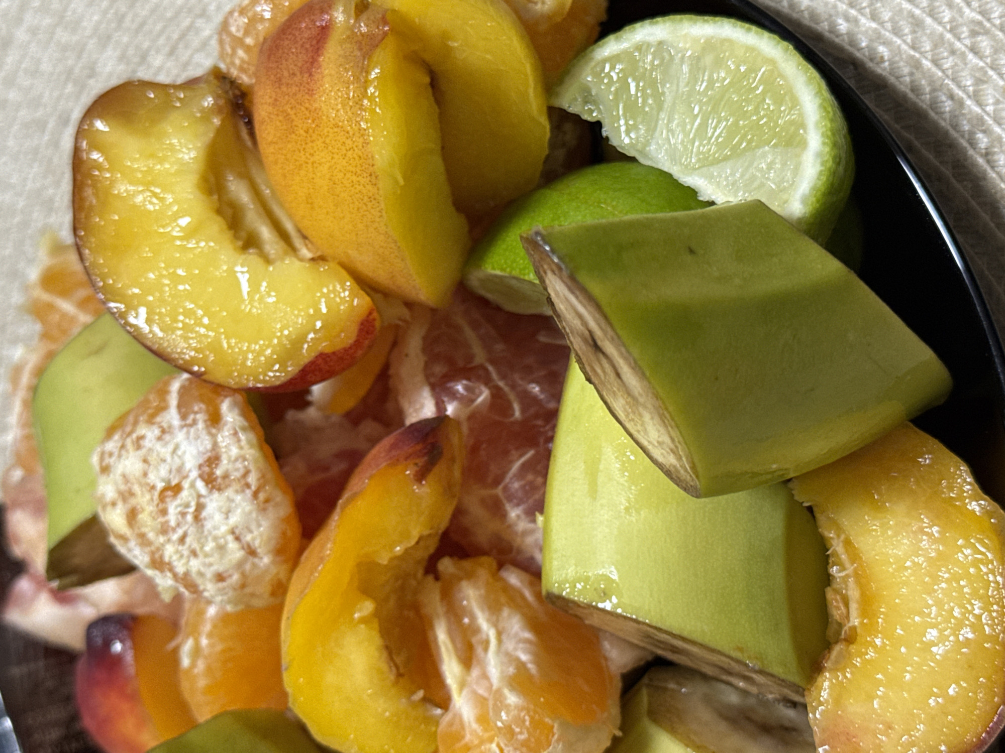 The image displays a close-up, high-angle perspective of a variety of cut fruits arranged on a black, smooth-surfaced platter. According to the prompt, the location is the city of Chaikovsky, Russia. The platter rests upon a light-colored, woven mat or placemat, visible in the lower right quadrant. The subjects primarily consist of segmented and sliced fruits. Prominently visible are multiple yellow-orange slices of peaches or nectarines, some exhibiting remnants of reddish skin and a glossy, moist surface. Interspersed among these are several peeled segments of mandarins or oranges, characterized by their distinct internal segmentation and visible white pith. Also present are multiple thick, light green pieces of fruit, consistent with unripe bananas or a type of melon, displaying minor brownish discoloration along their cut edges. At least two wedge-shaped slices of lime are also visible, showcasing green rind and segmented pale green pulp; one lime segment features an irregular cut. The composition indicates a prepared fruit assortment. No individuals are depicted, and no specific actions or interactions beyond the fruit arrangement are observable. The environment suggests an indoor setting, likely a dining or preparation area.
