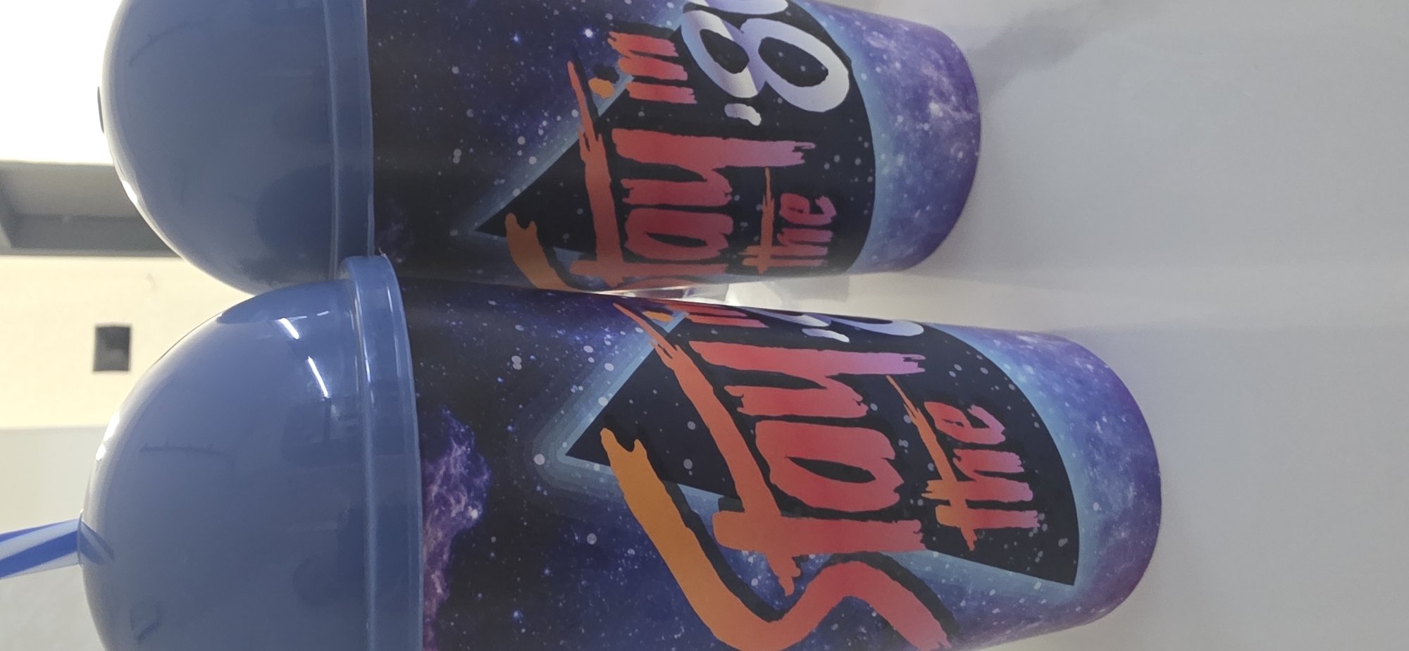 Two blue plastic cups with lids and straws sit on a white surface. The cups have a dark blue background with white speckles, resembling a starry sky. A purple and blue gradient triangle is visible behind text. On the left cup, the text "Stay the" is in bright red and orange lettering. On the right cup, the text "Stay in '80" is visible, with the "80" being in white lettering. Both cups are identical in design. The background is out of focus, showing a white wall and a dark rectangular object. The scene appears to be indoors, possibly in a cafe or shop. There are no people or animals in the image. The image does not contain any information about the city of Sumgayit, Azerbaijan.