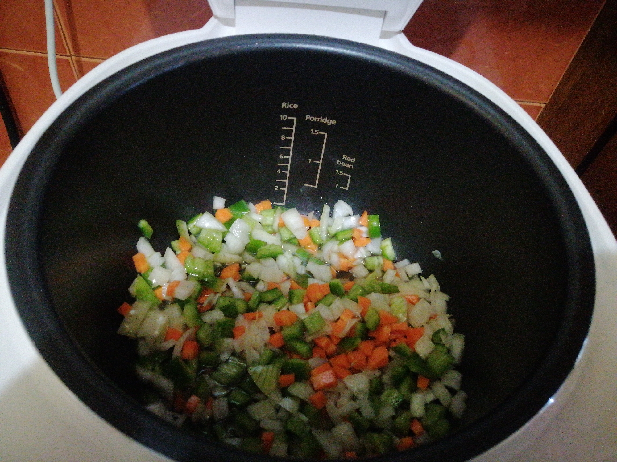 A rice cooker with diced vegetables inside sits on a brown tiled surface. The vegetables appear to be a mixture of white onions, green bell peppers, and orange carrots. The rice cooker is open, revealing a black inner pot with measurement markings for rice, porridge, and red beans. A white electrical cord is visible on the left side of the image. The location is not discernible from the image.