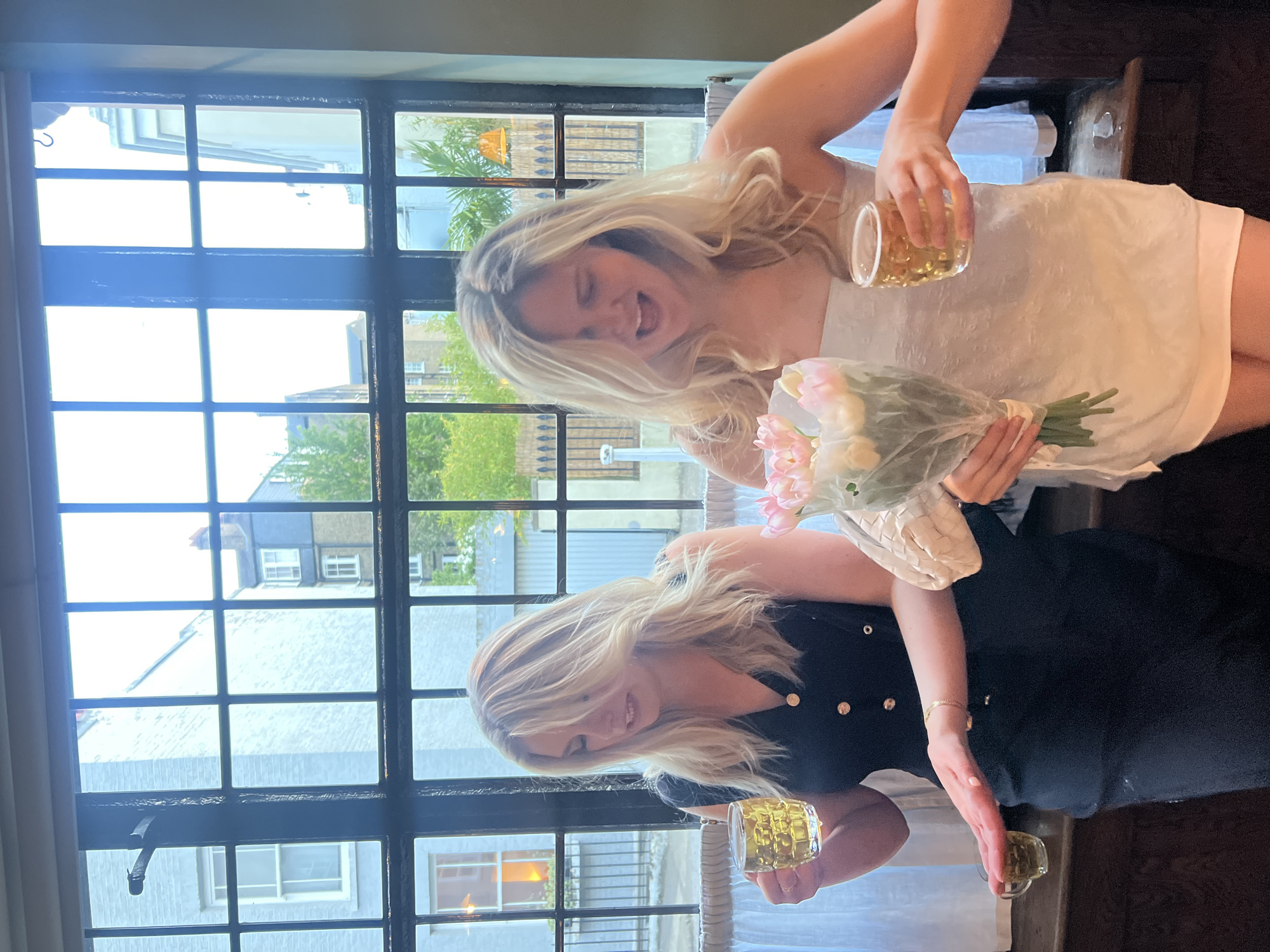 Two light-skinned women with blonde hair are standing in front of a window in an indoor setting, likely a restaurant or pub, based on the decor. The woman on the upper side of the frame is wearing a white dress, holding a bouquet of pink flowers and a glass with a yellow liquid. She is smiling with her mouth open. The second woman is wearing a black dress with gold buttons, holding two glasses containing yellow liquid and slightly smiling.
The window behind them is divided into multiple panes, and through it, the exterior view reveals buildings typical of London, United Kingdom, suggesting that the location may be in the city. The exterior architecture visible through the window is characterized by multiple floors, brick or stucco facades, and a mix of residential and commercial properties. The natural light suggests it is daytime.