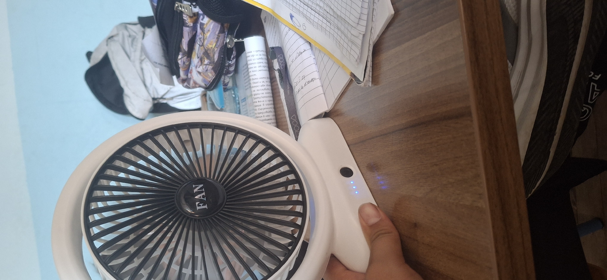 A close-up, eye-level shot captures a white portable fan placed on a wooden desk. The fan, a prominent object, features a black grill with the word "FAN" in white lettering at its center. To the right of the fan, a stack of open notebooks and papers are scattered across the desk, with a yellow ruler lying on top of one notebook. Further in the background, a partially visible backpack and other items are present. The desk surface appears to be made of polished wood with a visible grain. The lighting suggests an indoor setting, possibly in a home or office. The background wall is a light blue. The bottom left corner of the image shows a hand, with fingers extended and resting on the desk near the fan's base, which has four blue indicator lights.