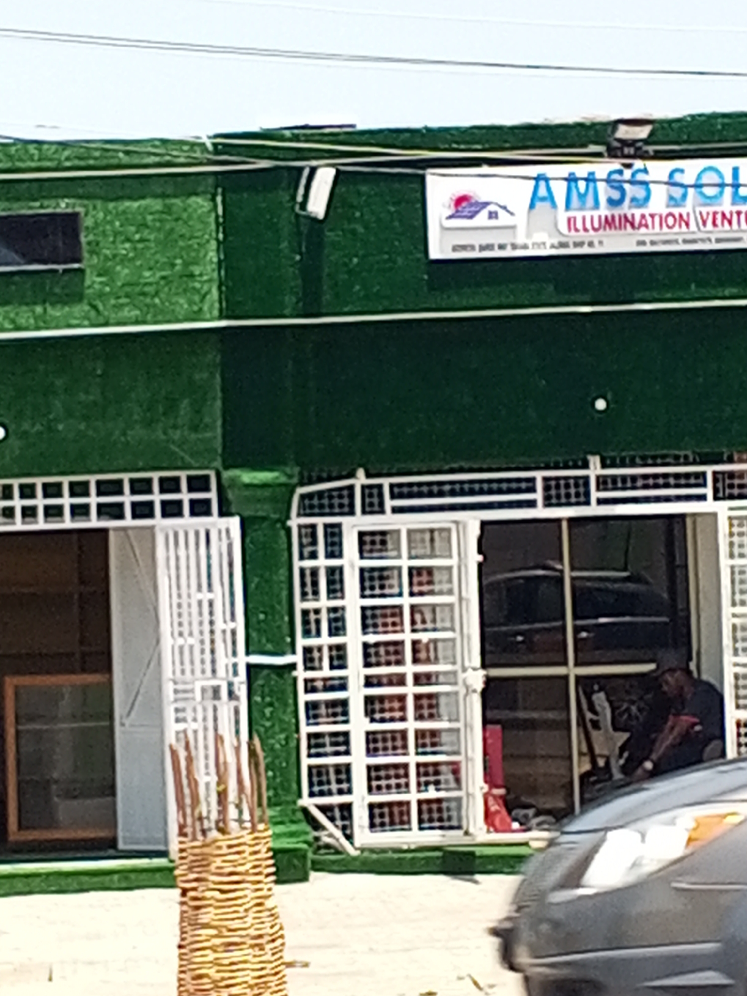 A man wearing a dark t-shirt, a red stripe on the sleeve, and a dark cap sits behind a counter inside a building with a green, textured exterior. The building has large glass windows with white grates. A dark-colored car is parked in front of the building, partially obscuring the view. A sign above the entrance reads "AMSS SOL ILLUMINATION VENTURES". The scene appears to be in an urban setting, possibly Jalingo, Nigeria.