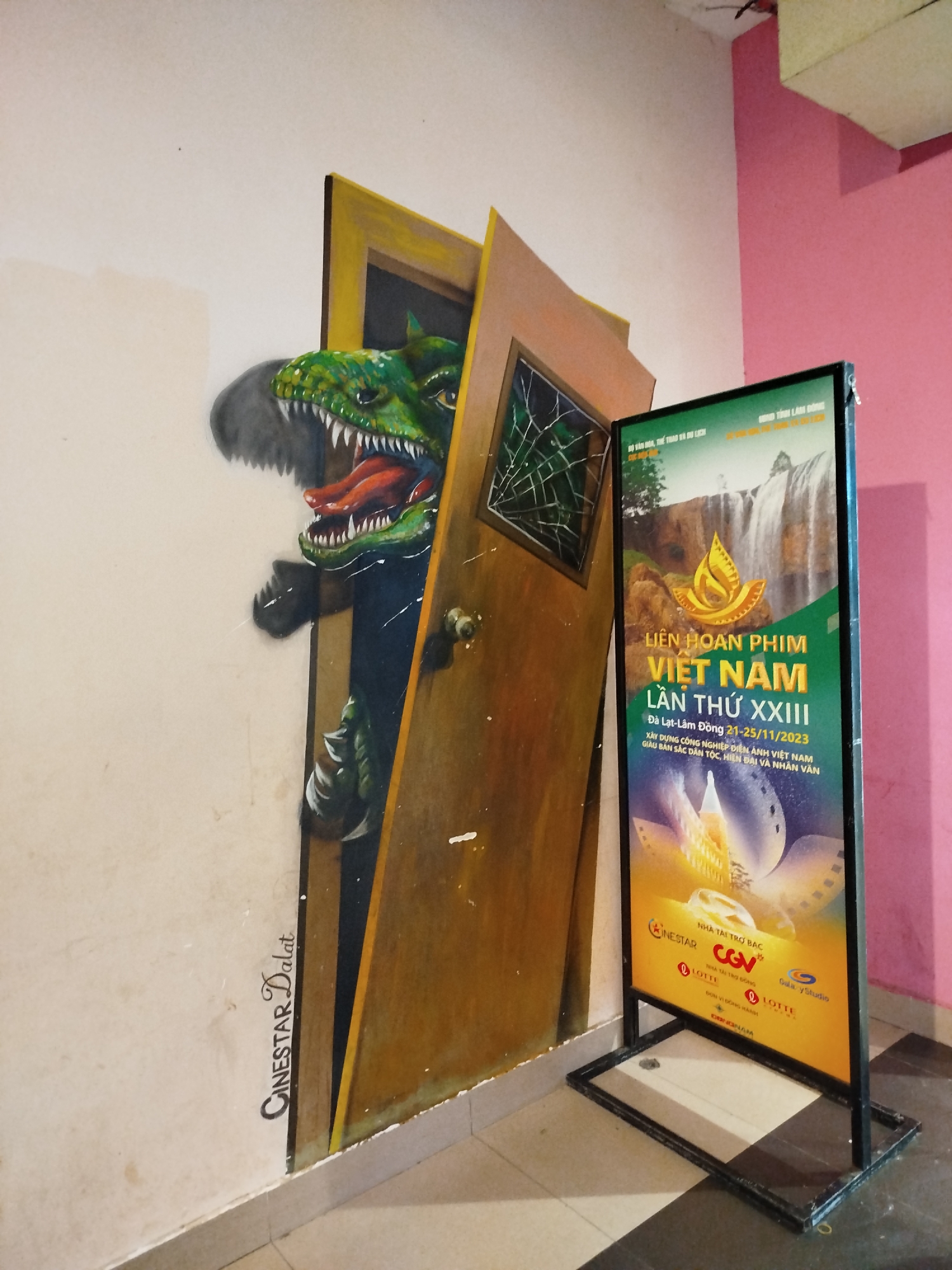 A room corner has a wall mural of a dinosaur emerging from a broken door, juxtaposed with a freestanding sign advertising the Vietnam Film Festival XXIII in Da Lat-Lam Dong from November 21-25, 2023. The mural includes the logo "CINESTAR Dalat." The sign also features logos of sponsoring companies, including CineStar, CGV, and Lotte. The floor is tiled in white and gray. The wall adjacent to the mural is pink.