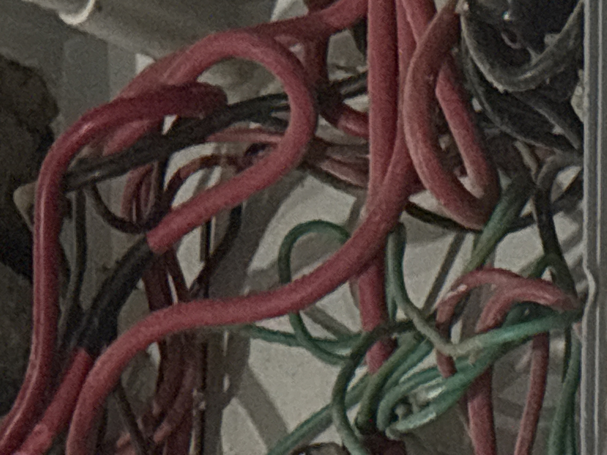 The image displays a close-up, static shot of a dense tangle of electrical wires. The primary subjects are numerous red-sheathed wires, heavily intertwined with several green and dark/black wires. The wires appear to be of various gauges and are coiled and looped irregularly, suggesting a chaotic or exposed wiring installation rather than a neatly organized system.
The background consists of muted gray and off-white tones, possibly indicating a wall or the inside of an electrical box or conduit, with subtle textures and shadows adding to the depth of the scene. No specific activity is taking place in the image; it is a snapshot of the wires as they are.
Notable details include the clear distinction between the red, green, and dark wire colors. The image has a visible graininess or noise, hinting at potentially low-light conditions or a camera with limited resolution. There are no people, explicit emotions, time of day indicators, weather conditions, or visible text within the frame. The location context of Angwan Sayawa, Nigeria, is not visually discernible from the image itself.
