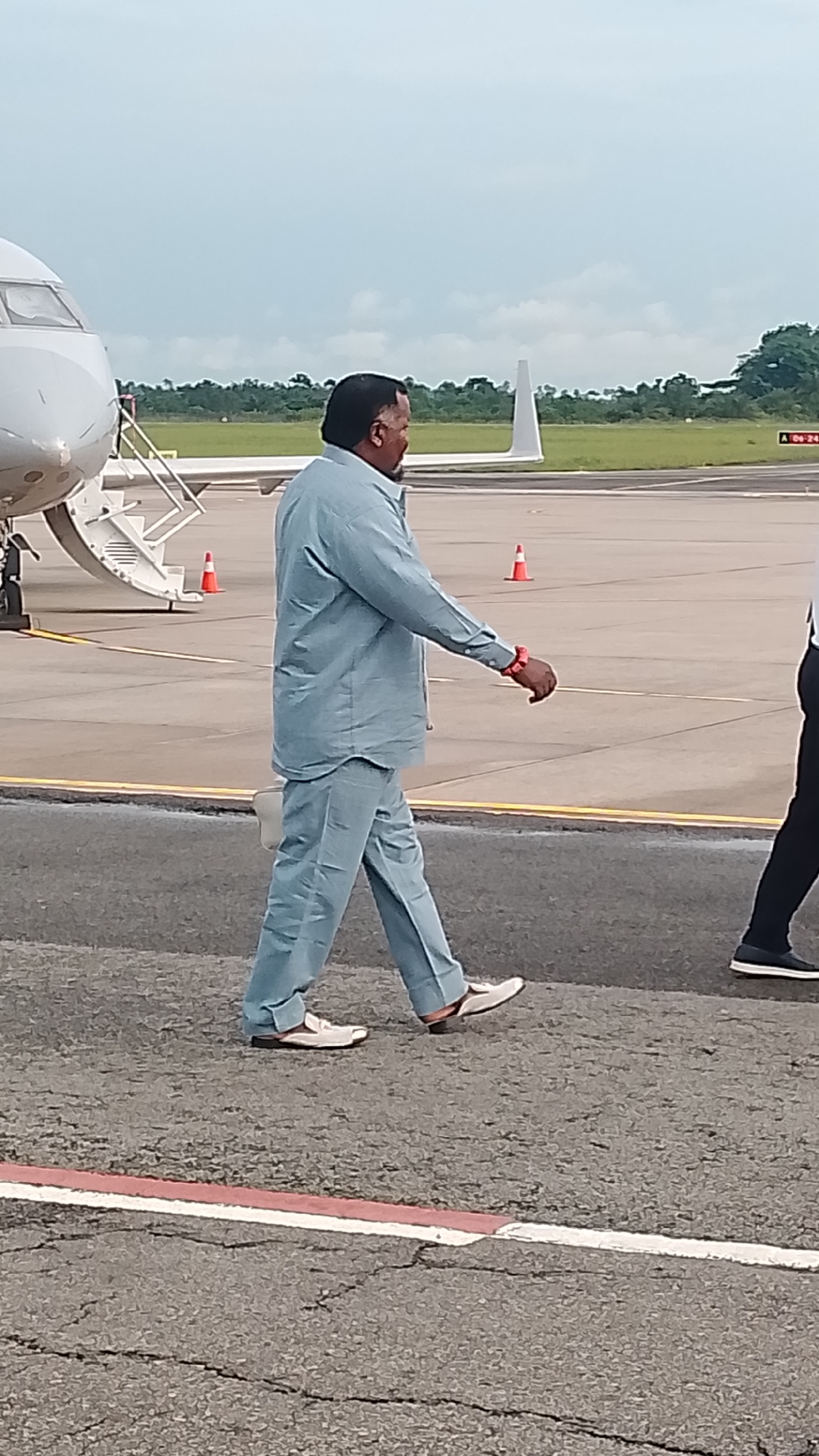A man in a light blue suit and white shoes walks across an airport tarmac. He is walking from left to right, with his body angled towards the right side of the frame. The man has dark hair and a beard. He is wearing a red wristband on his left wrist. Behind him, a portion of a white airplane with stairs is visible on the left. Orange traffic cones are scattered across the tarmac. In the background, there are trees and a cloudy sky. The location is identified as the airport in Orerokpe, Nigeria.
