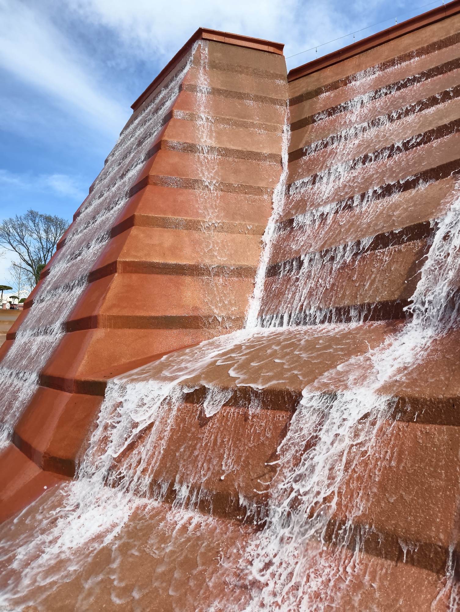 This image captures a large, tiered waterfall structure, resembling a stepped pyramid or ziggurat, in an outdoor setting in Krasnodar, Russia. The structure is constructed from a reddish-brown material, possibly concrete or stone, and features multiple receding steps. Water actively cascades down these steps, creating frothy white streams and ripples as it flows from one level to the next.
The perspective is low, looking up towards the top of the waterfall feature, which fills a significant portion of the frame. The left side of the structure shows more of its receding, angular design, while the right side focuses on the intricate flow of water at closer range.
In the upper background, a bright blue sky with wispy white clouds is visible, indicating a clear, possibly sunny day. To the far left, the bare branches of a tree are visible, suggesting it might be winter or early spring. No people or visible text are present in the image. The scene conveys a sense of architectural grandeur and the dynamic movement of water within a public park or landscape.