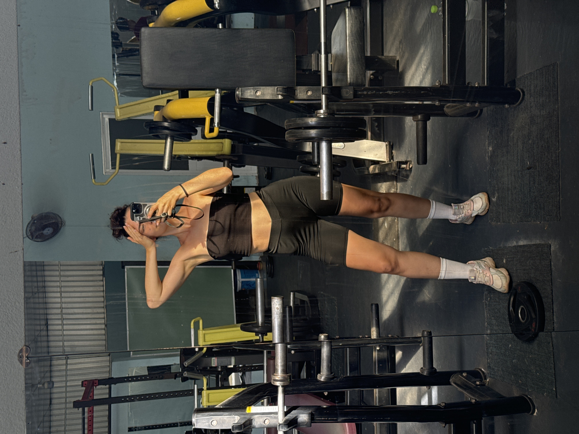 The image shows a person standing in a gym in Mũi Né, Vietnam, taking a selfie in a mirror. The person is wearing a black sports bra and black shorts with white socks and athletic shoes. The gym equipment is visible, including weight machines with yellow and black components and plates on the floor. The floor appears to be a dark, likely rubberized material. The reflection in the mirror reveals more gym equipment and a light blue wall with a fan.