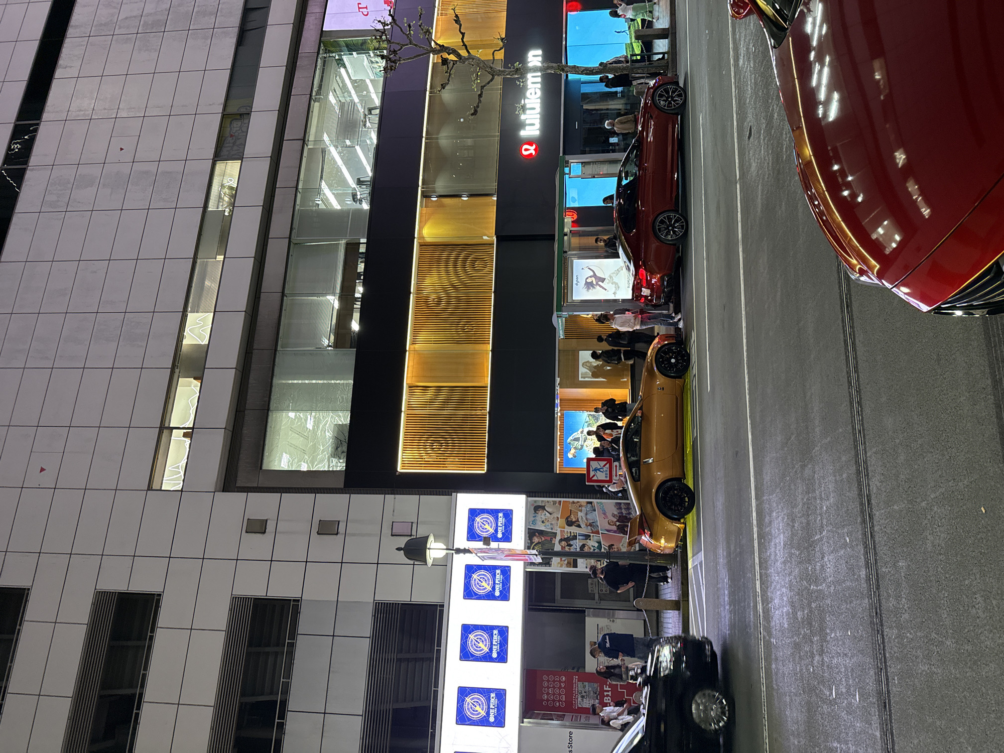 The image depicts an urban street scene at night, likely in Shibuya, Japan. Visible are: a multi-story building with the "lululemon" logo, several parked cars (one red, one gold, and one black), and advertisements. Pedestrians are seen near the storefront, engaging in activities such as looking at merchandise and standing nearby. A series of vertical advertisements featuring the "ONE PIECE" franchise are also displayed. The lighting is a mix of building lights, advertisement lights, and car headlights.
