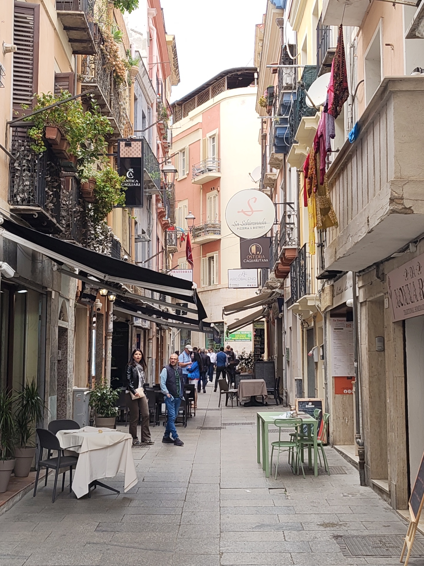 Here is a detailed description of the image:
The image captures a narrow street in Cagliari, Italy, lined with buildings of varying heights and architectural styles. The street, paved with light-colored stone blocks, features outdoor seating areas for restaurants and bistros.
On the left side of the street, several establishments have awnings extending over their facades, providing shade to the outdoor tables. Balconies with ornate iron railings are visible above the storefronts, adorned with greenery and hanging plants. Signs for "ANTICA CAGLIARI" are displayed on a building.
On the right side, colorful fabrics and clothing hang from balconies, adding a touch of vibrancy to the scene. A sign reads "Sa Schironada PIZZERIA & BISTROT".
Several people are walking through the street, some appearing to be tourists or locals enjoying a stroll. A couple stands near a table, with the woman in a leather jacket and the man in a vest. Other groups of people are seen further down the street, suggesting a bustling atmosphere.
The overall impression is of a lively, pedestrian-friendly street in Cagliari, characterized by its mix of architectural styles, outdoor dining options, and bustling activity.