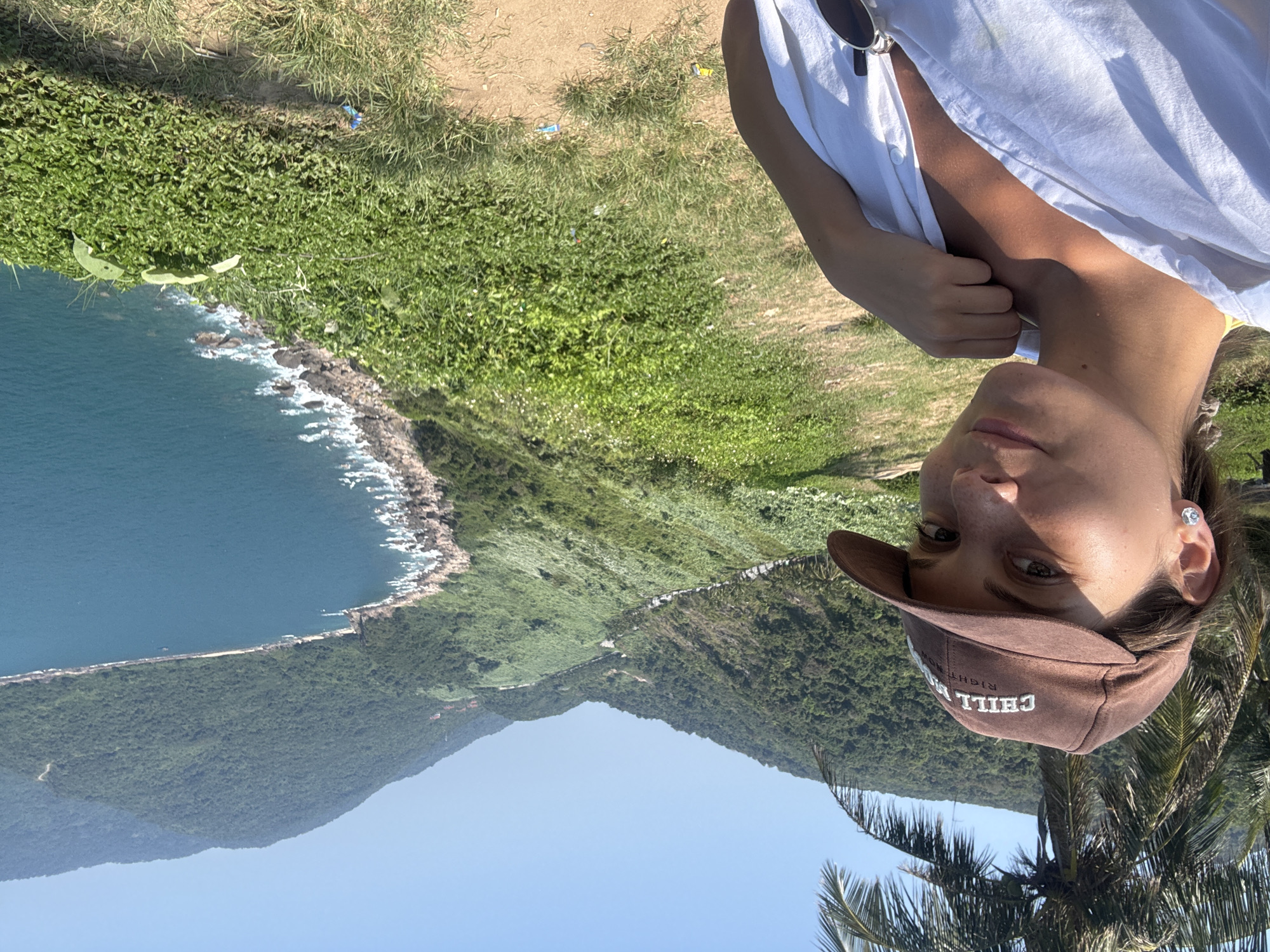 A person stands on a grassy hill overlooking a scenic coastline and mountainous landscape in Đà Nẵng, Vietnam. The person, wearing a white shirt and a brown cap with the word "CHILL" embroidered on it, gazes directly at the camera. Behind them, the bay stretches out, with clear turquoise water meeting a rocky shoreline and lush greenery. Palm trees are visible in the foreground. A road snakes along the coast.