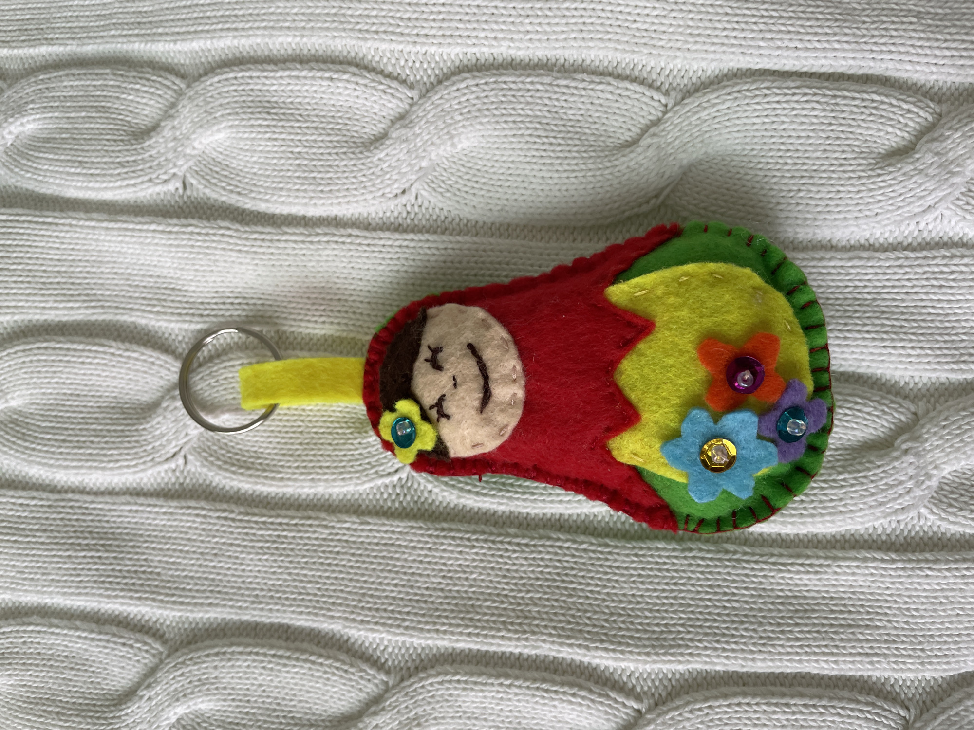 A handmade felt keychain resembling a Russian doll lies on a white cable-knit sweater. The doll has a red body, a cream-colored face with embroidered features, a yellow loop for the keychain ring, and colorful felt flower embellishments. It is photographed indoors. No location is recognizable in this scene. Лесной Пейзаж Кп (шарапово, Russia is not visible.