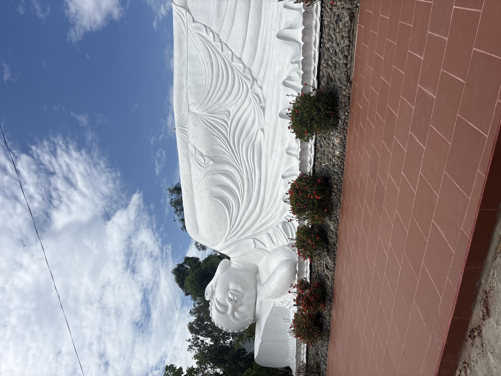 This image captures a massive white reclining Buddha statue, a prominent religious monument located in Cam Ranh, Vietnam. The statue is depicted lying horizontally, with its serene head, featuring closed eyes and elongated earlobes, resting on its right arm on the left side of the frame. Its body is draped in robes intricately carved with flowing, wave-like patterns, adding texture and detail to the brilliant white surface.
The Buddha statue rests on a base made of dark, rough stones. In the foreground, a reddish-brown tiled pathway, composed of rectangular blocks, stretches across the right side of the image. Between the pathway and the statue's base, a narrow strip of dark pebbles is visible, interspersed with several green bushes adorned with clusters of vibrant red flowers.
Above the statue, a bright blue sky fills the upper portion of the scene, dotted with scattered white clouds, suggesting clear, sunny weather, likely during the day. Lush green trees and foliage are visible behind and partially above the statue, providing a natural backdrop and indicating a verdant, possibly hilly, environment. A thin black wire traverses the sky in the upper left background. The overall scene conveys a sense of peace and tranquility, characteristic of a sacred site. No human activity or visible text is present.