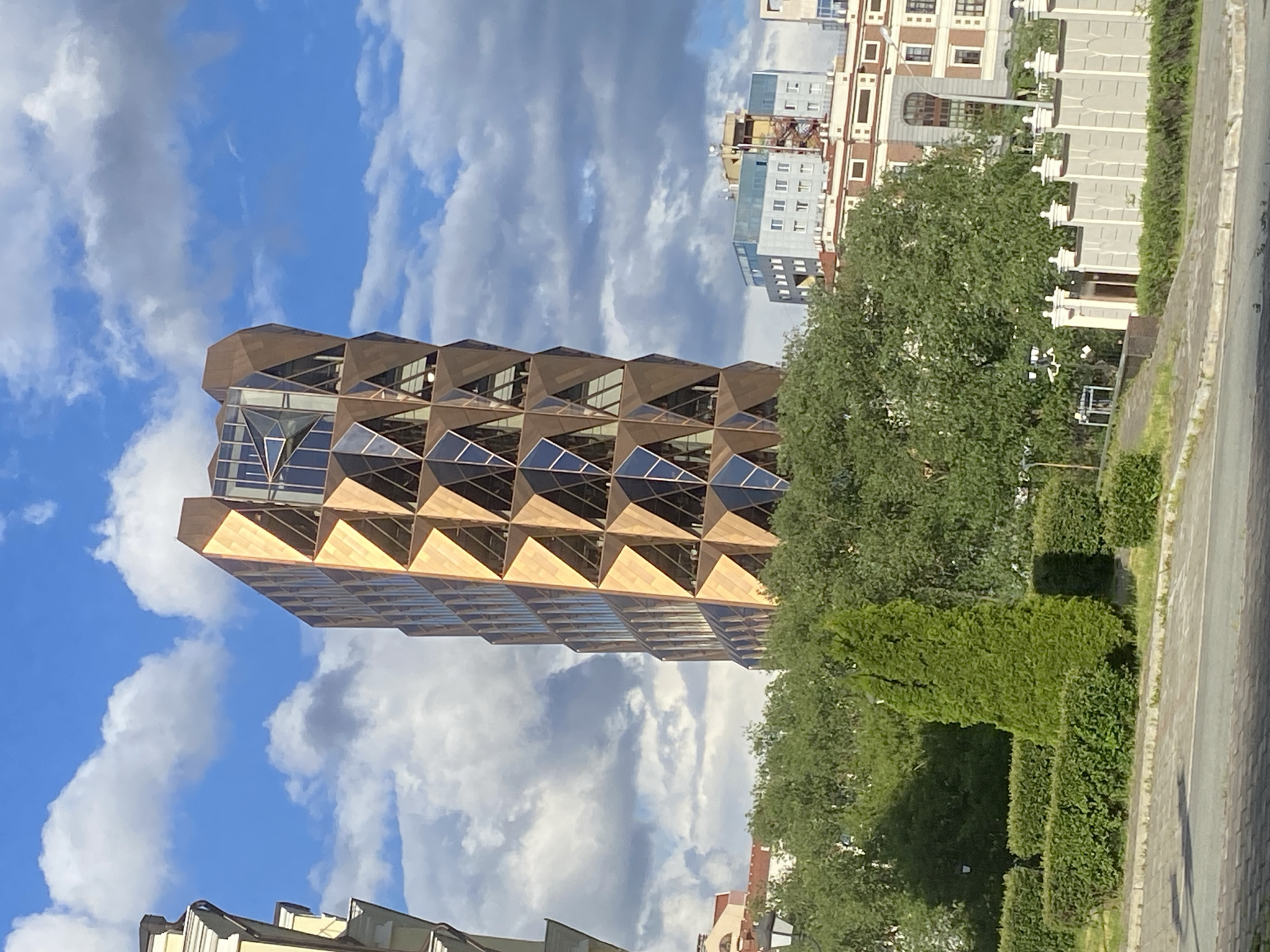 The image showcases a distinctive architectural structure, identified as the "Pineapple Building" located in Yekaterinburg, Russia. The building, characterized by its pyramidal patterns on the facade, is positioned against a backdrop of a partly cloudy sky. Adjacent to the building is an area of greenery, including trees and manicured hedges, running parallel to a paved road. In the background, other buildings with various architectural styles can be observed, typical of a cityscape.