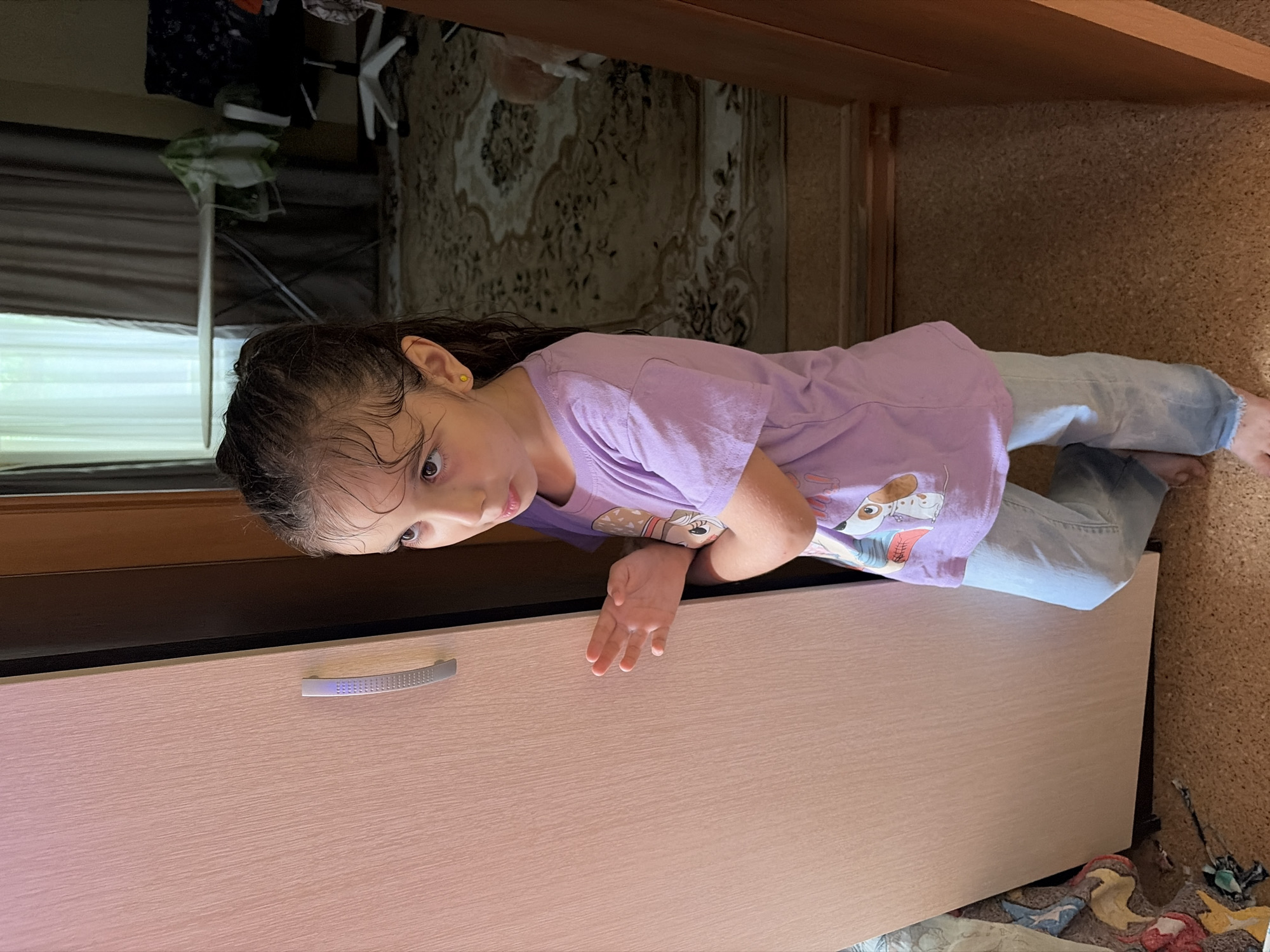 A young girl with dark, wet hair and brown eyes is standing by a light-colored wardrobe in an indoor setting. She is wearing a purple t-shirt with a graphic design of dogs and light blue jeans. Her right hand is raised, with her fingers slightly spread. The background includes a doorway, a rug with a patterned design, an ironing board, and an office chair. The lighting suggests it is daytime, with light coming from a window through sheer curtains. The scene appears to be a residential interior, potentially a bedroom or hallway. There is no specific indication of the city of Tambov, Russia.