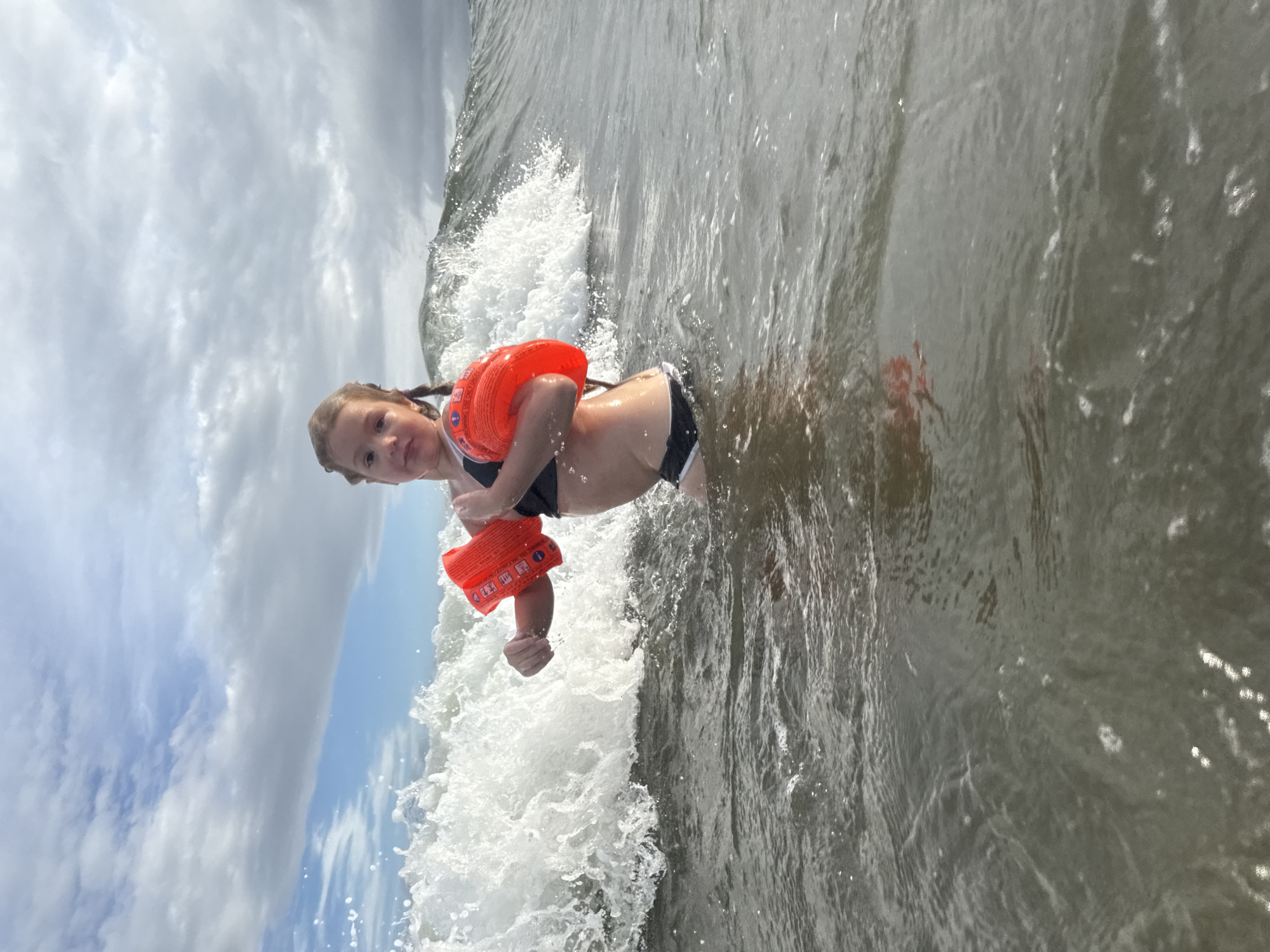 The image shows a young girl with blonde braided hair, wearing a black swimsuit and an orange flotation device, in the ocean. She is partially submerged in the water, with a wave breaking around her. The sky is cloudy, and the sun appears to be out, suggesting it might be daytime. The girl is looking at the camera with a playful expression. The water is choppy, and there are whitecaps from the breaking wave. The scene is likely a beach or coastal area. There is some text visible on the orange flotation device, but it is too small to read. The location is Vladivostok, Russia.