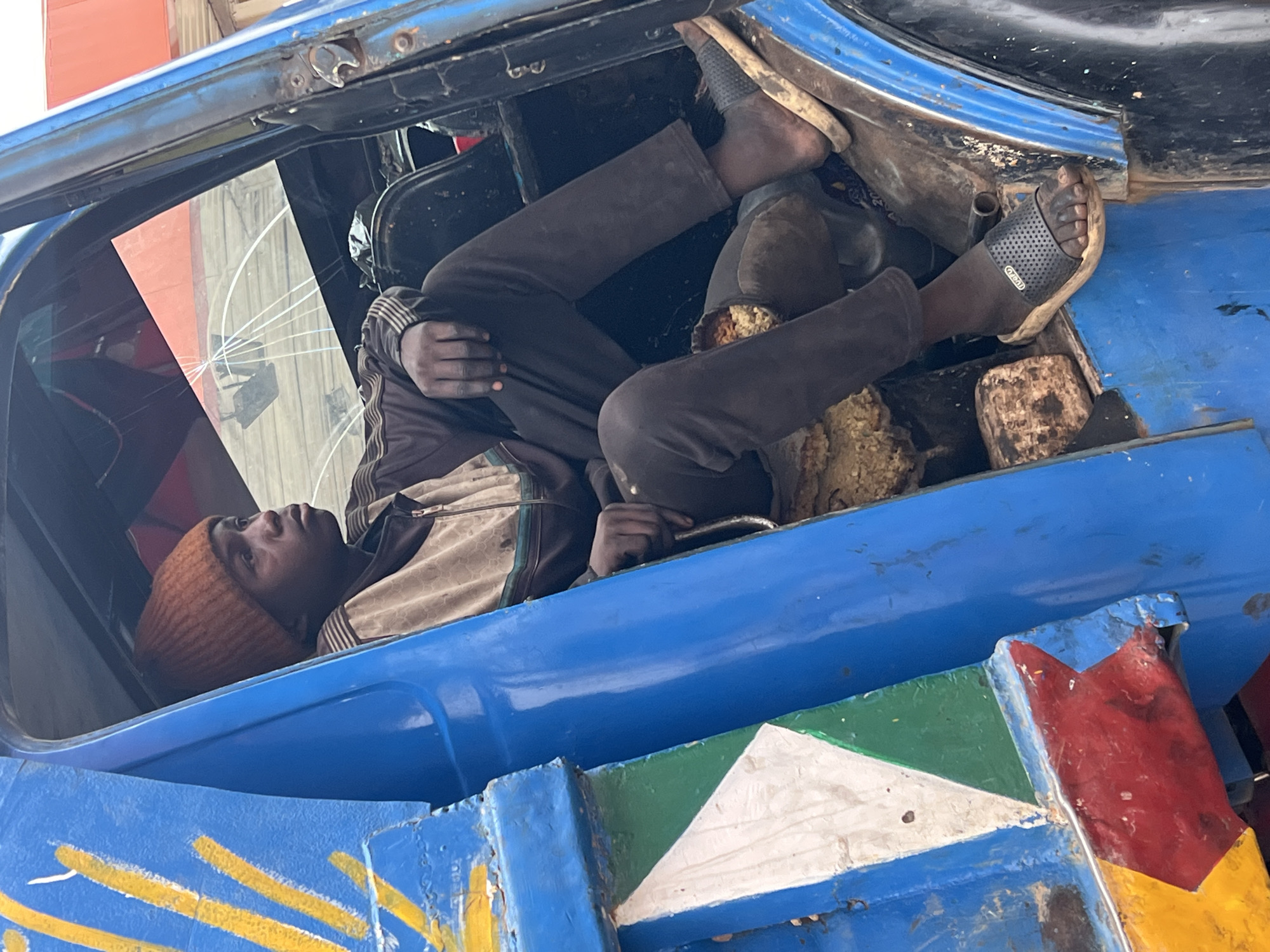 A person wearing a brown beanie, beige striped shirt, brown jacket, and brown pants is lying inside a blue painted vehicle. The person is also wearing black sandals. The vehicle has a cracked windshield and painted geometric patterns in red, green, yellow, and white. The location is Jos, Nigeria.