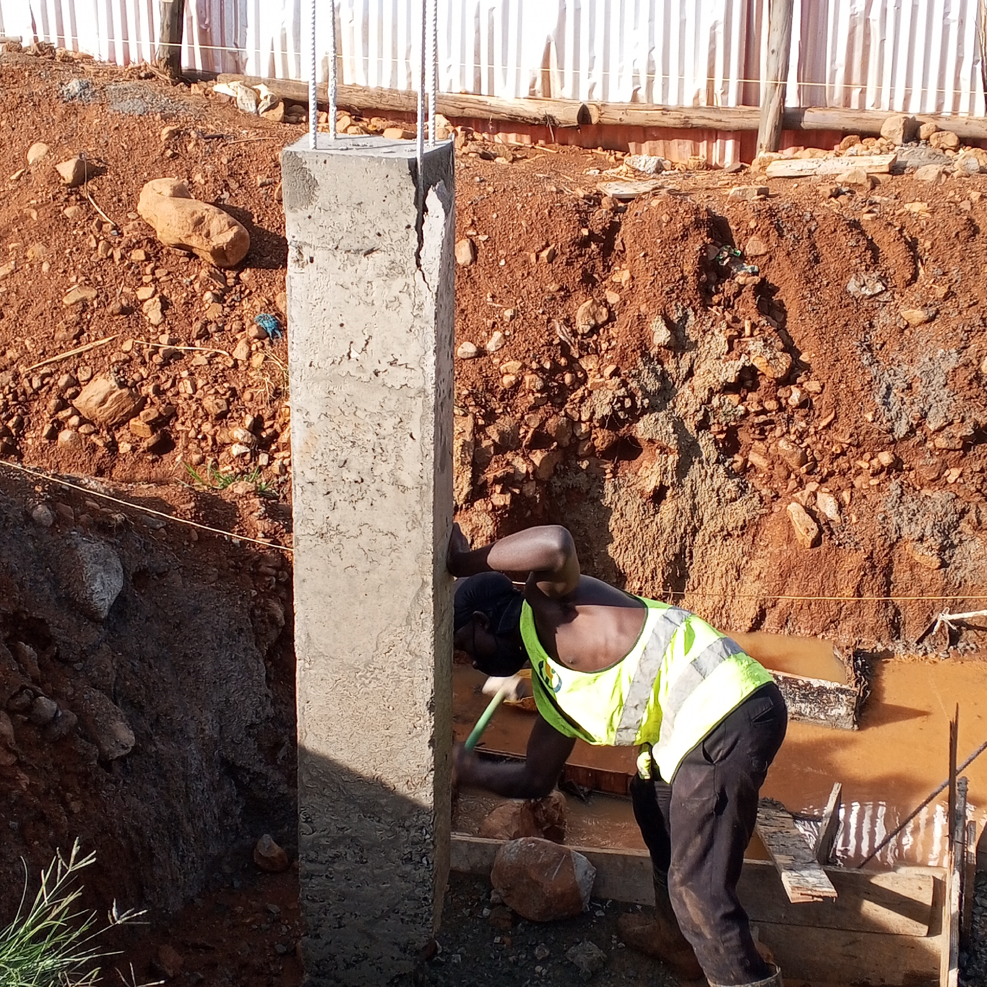 A construction worker in a bright yellow safety vest and black pants is working at a construction site in Borabu Chitago ward, Kenya. He is using a hammer or similar tool on the base of a tall, rectangular concrete column with exposed rebar extending from the top. The area around the column is excavated, revealing red-brown soil and rocks. There is standing water in the excavated area. A fence of corrugated metal sheets and wooden supports is visible in the background.