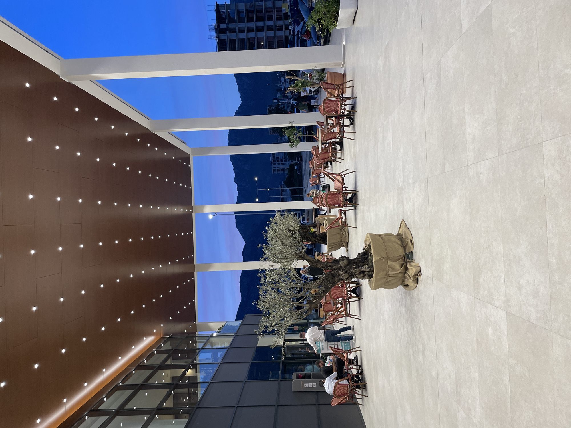 The image depicts an outdoor patio area in the city of Bar, Montenegro. The patio features white tiled flooring, red wicker chairs, and an olive tree potted in burlap. In the background, a mountain range is visible under a blue sky. To the left, a building with a brown facade and evenly spaced light fixtures is seen. The patio is framed by white pillars. Several people are present, including individuals seated at tables and a man in a white shirt walking past. Construction of another building can be seen in the background.